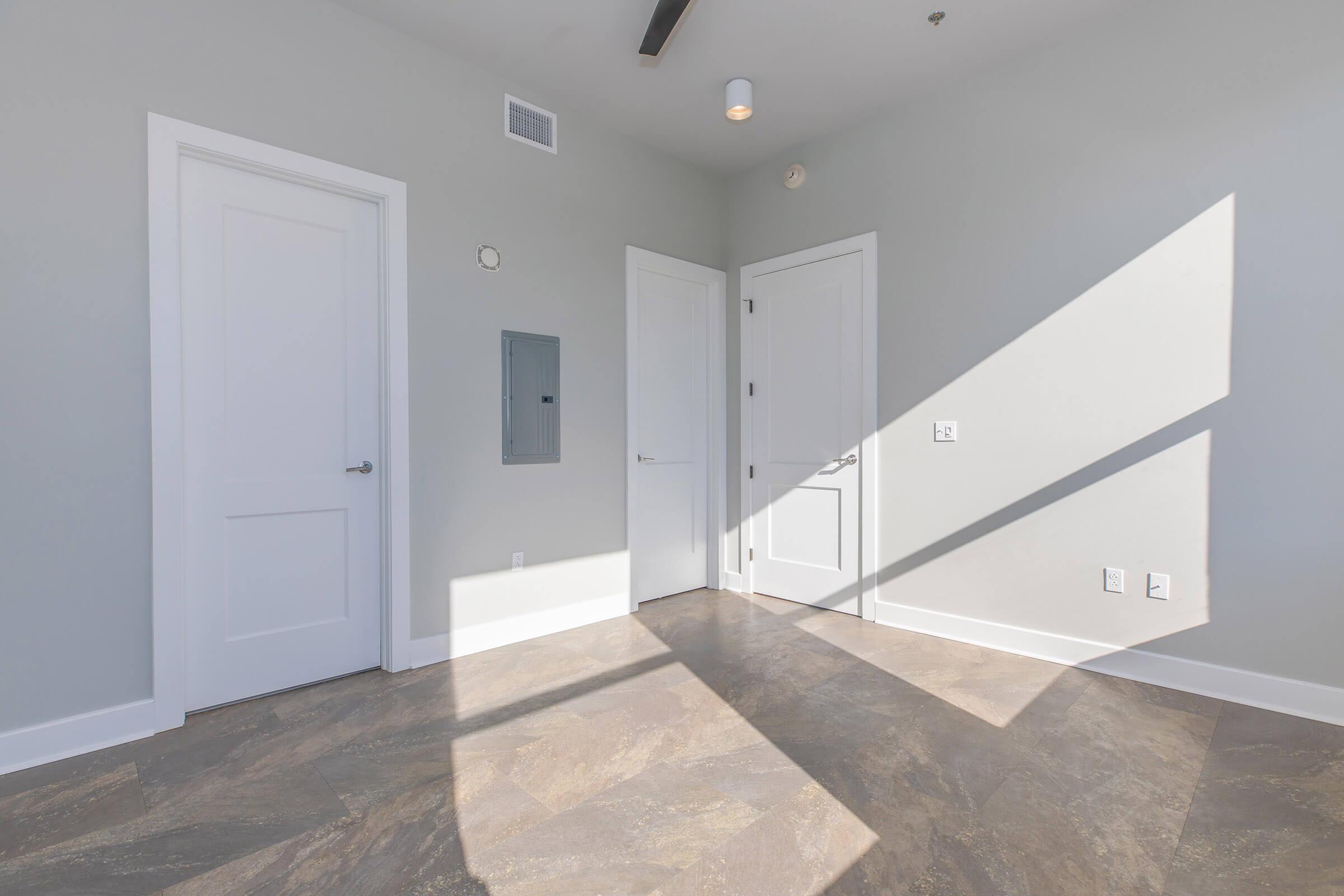 Empty room with light gray walls and a large floor area featuring beige tiles. There are three white doors—two are closed and one is slightly open. A light fixture is mounted on the ceiling, and shadows from the light create diagonal patterns on the floor. A mail slot is visible on the wall near the entrance.