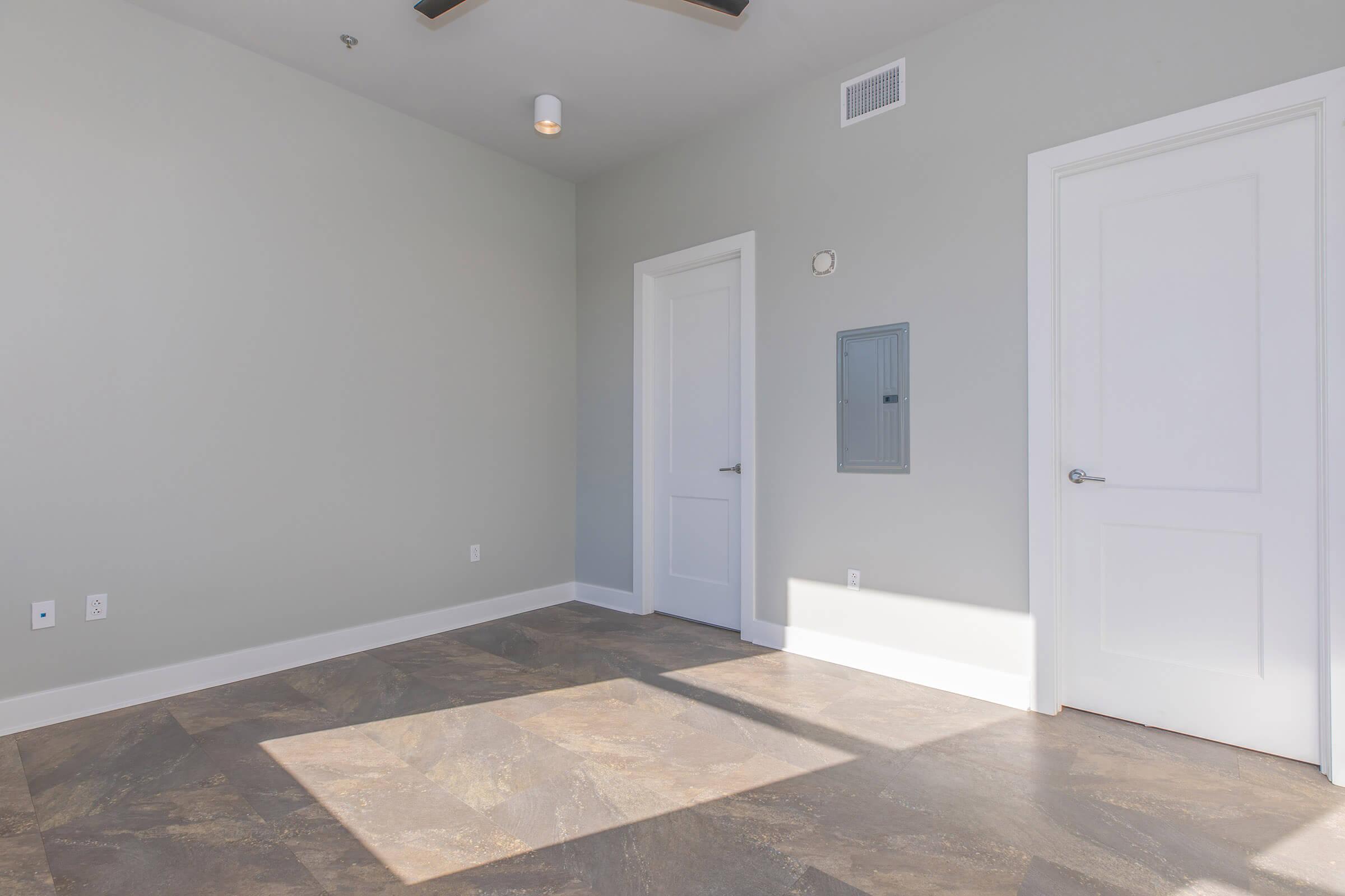 A minimally designed interior room with light gray walls and a large window casting sunlight onto the floor. There are two white doors, one leading to an adjacent space and the other being a closet or storage area. A ceiling fan is visible, and the flooring is dark, possibly tile or laminate.