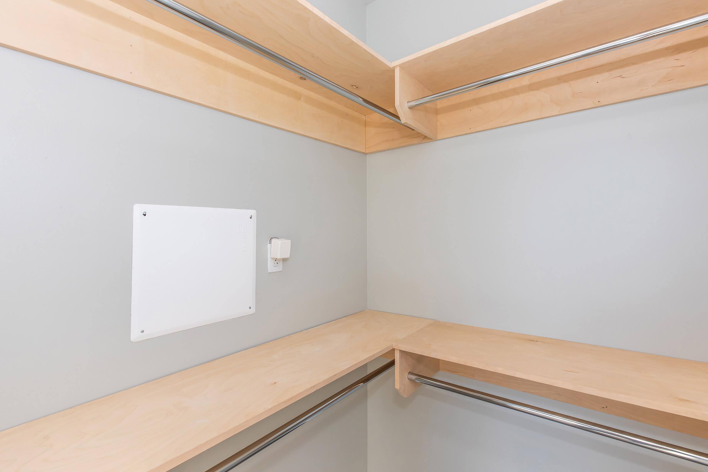 Empty closet with light gray walls and wooden shelves. There are two horizontal metal rods for hanging clothes, and a blank white panel on the wall, near a small electrical outlet. The overall design is minimalist and modern.