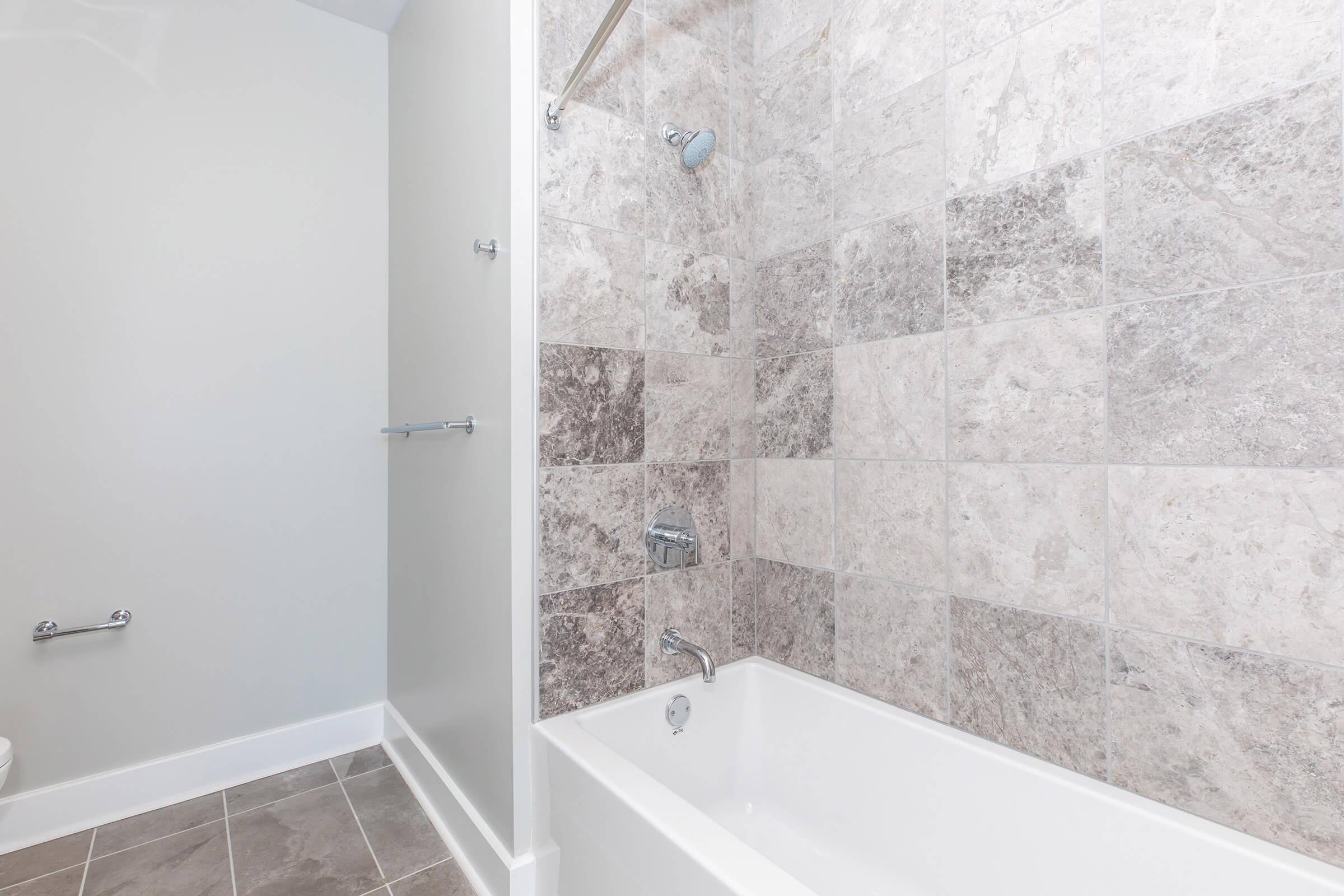 A modern bathroom featuring a clean, tiled shower area with gray and beige marble-like tiles. There is a bathtub with a chrome faucet and showerhead. The floor is tiled in a lighter gray, and there are wall-mounted grab bars. The walls are painted a light color, creating a spacious feel.