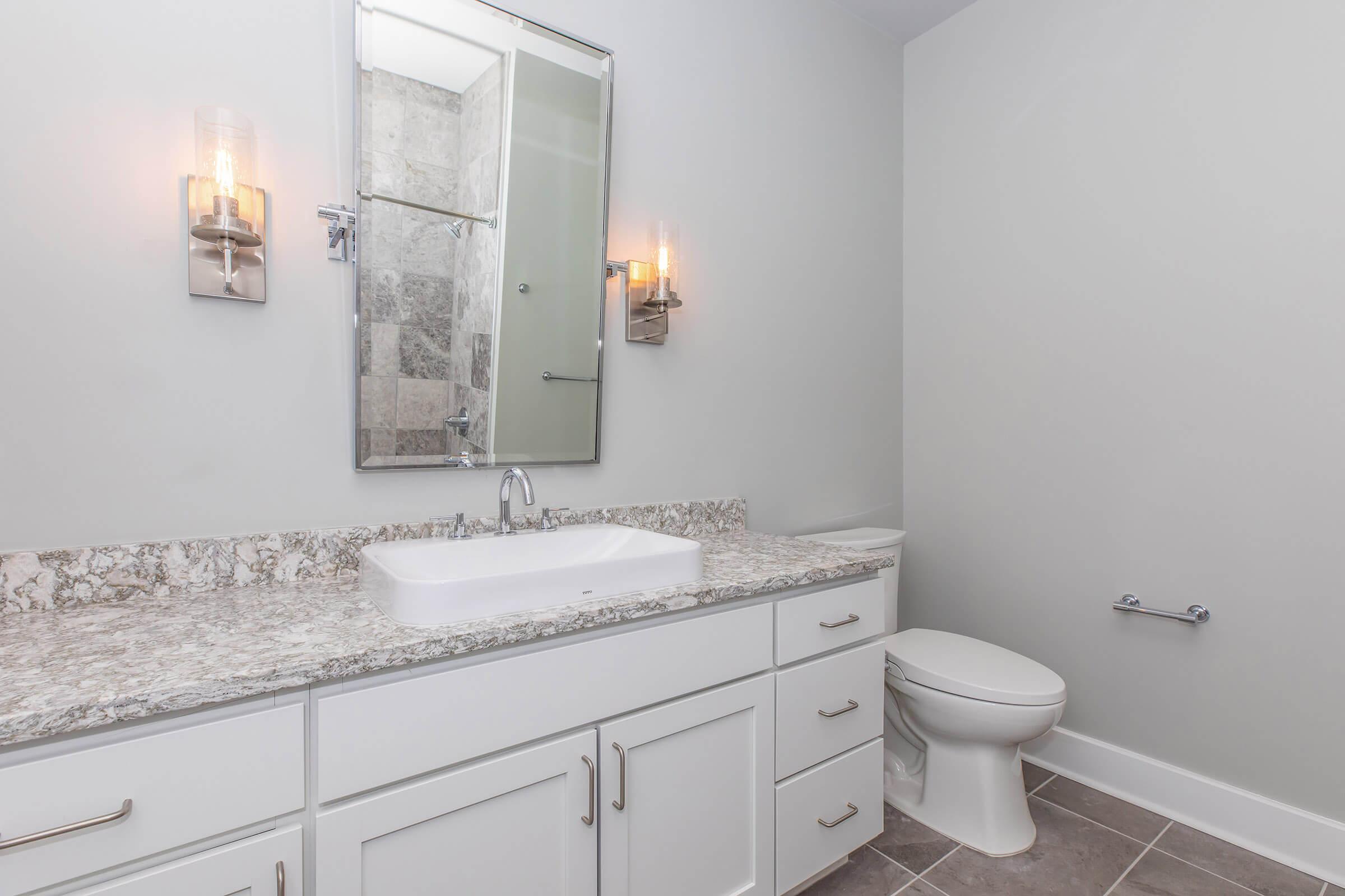 A modern bathroom featuring a white vanity with a rectangular sink, a large mirror above it, and decorative wall sconces. The walls are light gray, and there is a tiled shower in the background. A toilet is positioned to the right, with a toilet paper holder on the wall. The floor is tiled in a neutral shade.