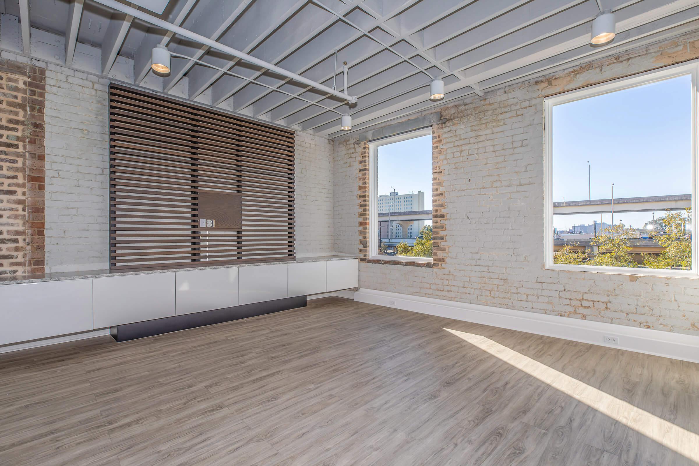 A spacious, modern room featuring exposed brick walls, large windows allowing natural light, and wooden flooring. The ceiling has a unique beam design, and there is a stylish wall fixture with horizontal wooden slats. The room appears empty and is designed with a contemporary aesthetic.