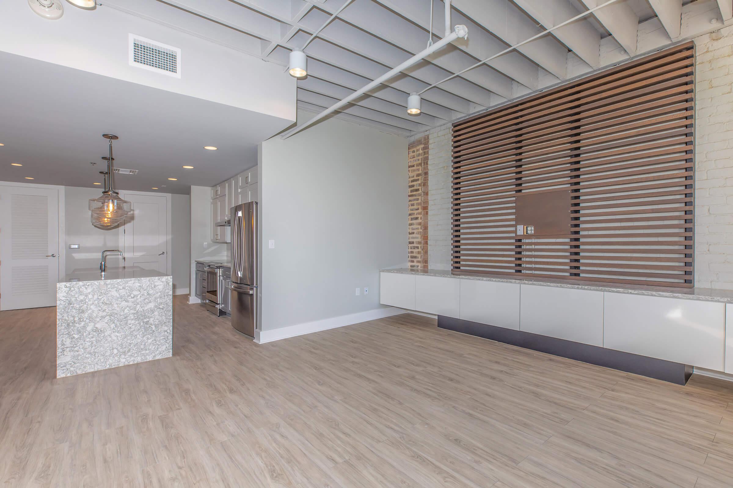 Modern, open-concept living area featuring a spacious kitchen with a large granite island, stainless steel appliances, and a stylish pendant light. The adjacent living space has wooden flooring and a light-colored wall with decorative wooden slats. Neutral tones create a bright and airy atmosphere.