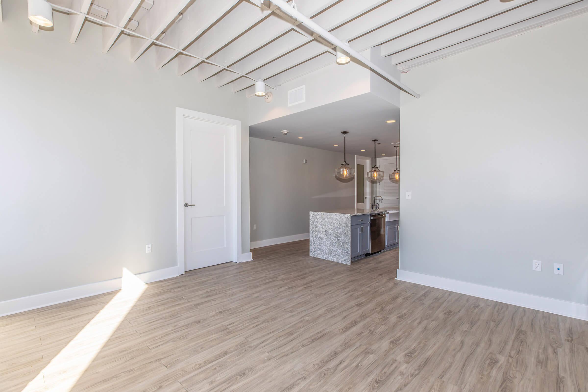 Spacious, modern living area with light wood flooring and high ceilings. A kitchen is visible in the background, featuring gray cabinetry and pendant lighting. The walls are painted a soft gray, and there is a doorway leading to another room. Natural light enters from the left, creating a bright atmosphere.