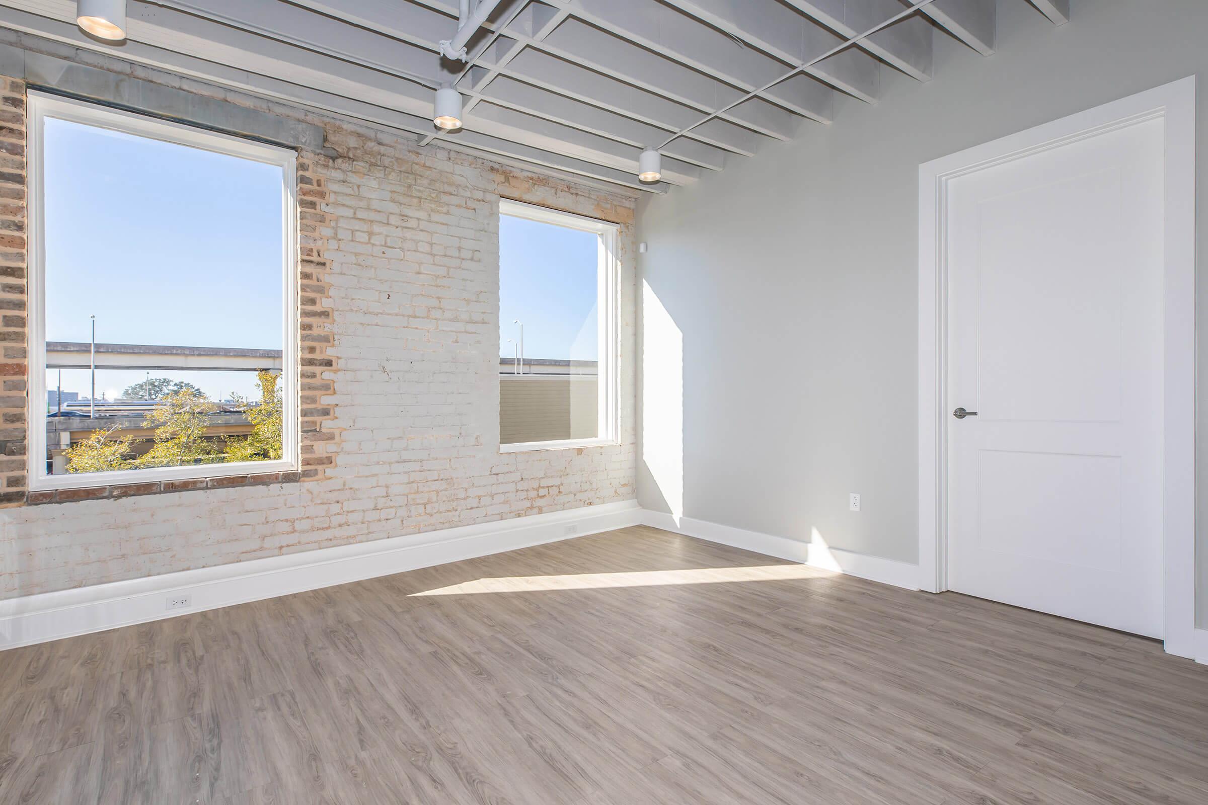 Bright, empty room with large windows allowing natural light to fill the space. The walls feature exposed brick and a smooth gray finish. The ceiling has a modern design with visible beams, and the floor is light-colored wood. A single white door leads to another area.