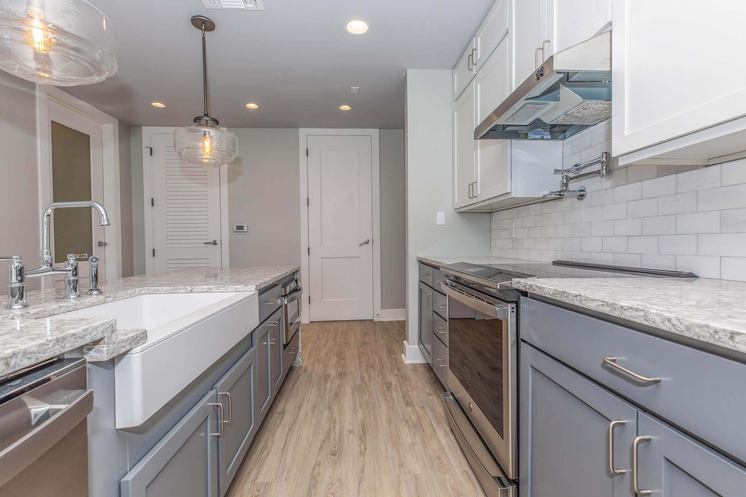 Modern kitchen featuring gray cabinetry, a large white farmhouse sink, stainless steel appliances including an oven and vent hood, and a stylish pendant light. The space has light-colored wood flooring and white subway tile backsplash, creating a clean and contemporary aesthetic.