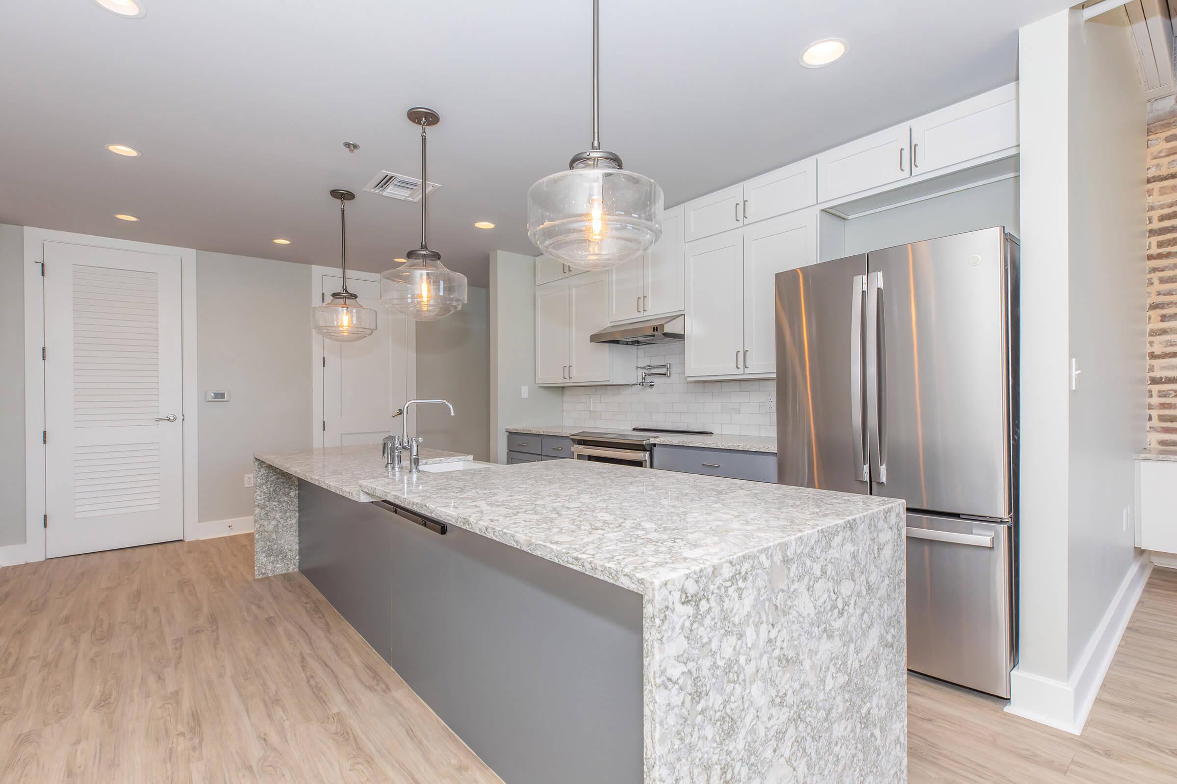 Modern kitchen featuring a large gray island with a marble countertop, stainless steel appliances, including a refrigerator and oven, and white cabinetry. The space is well-lit with pendant lights hanging above the island, and a light-colored wood floor complements the overall design.