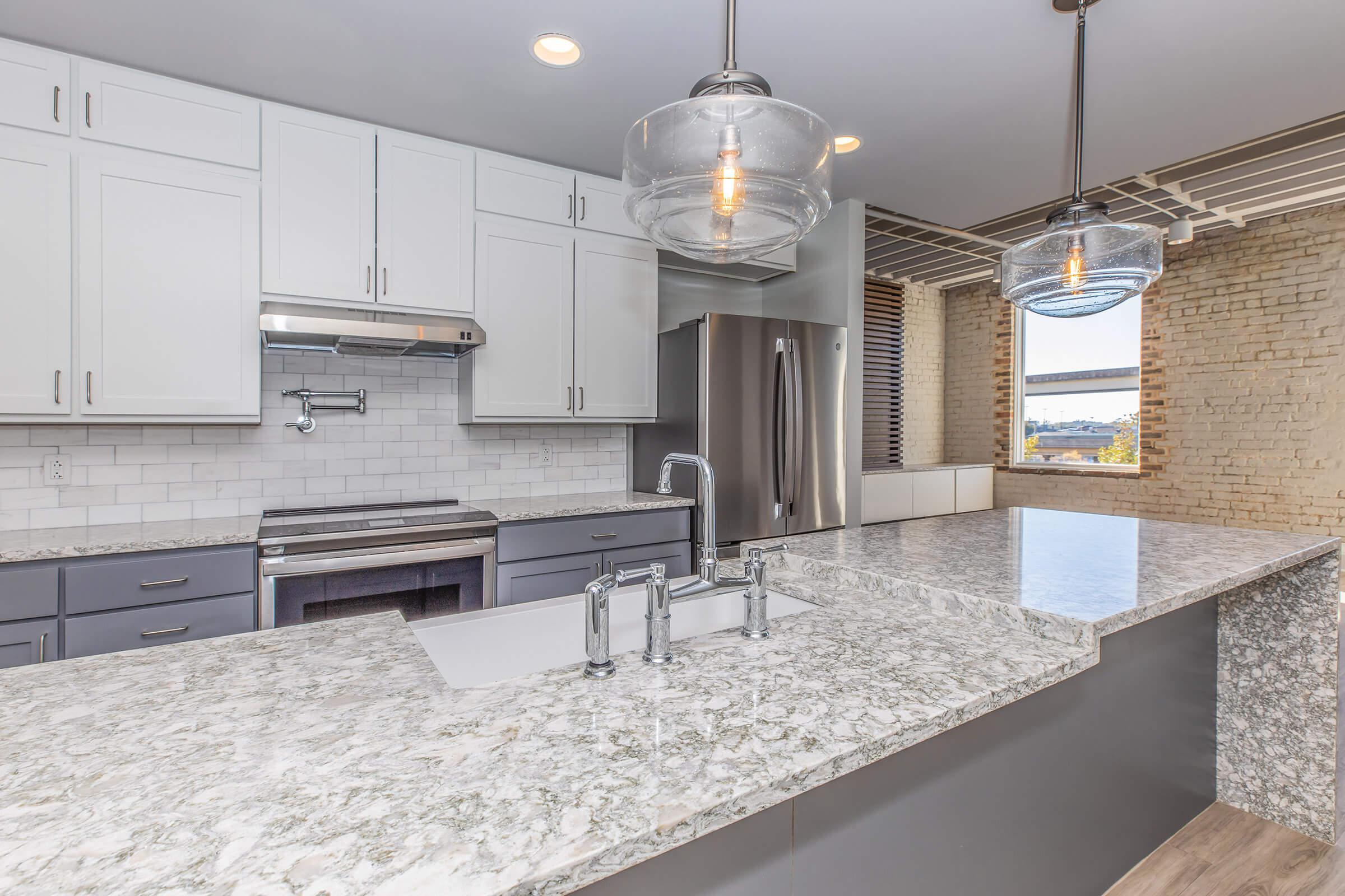 Modern kitchen featuring a large granite countertop island with a built-in sink, stainless steel appliances, and pendant lighting. The cabinetry is a mix of white and dark gray, and there are exposed brick walls, offering a sleek and spacious ambiance with natural light.