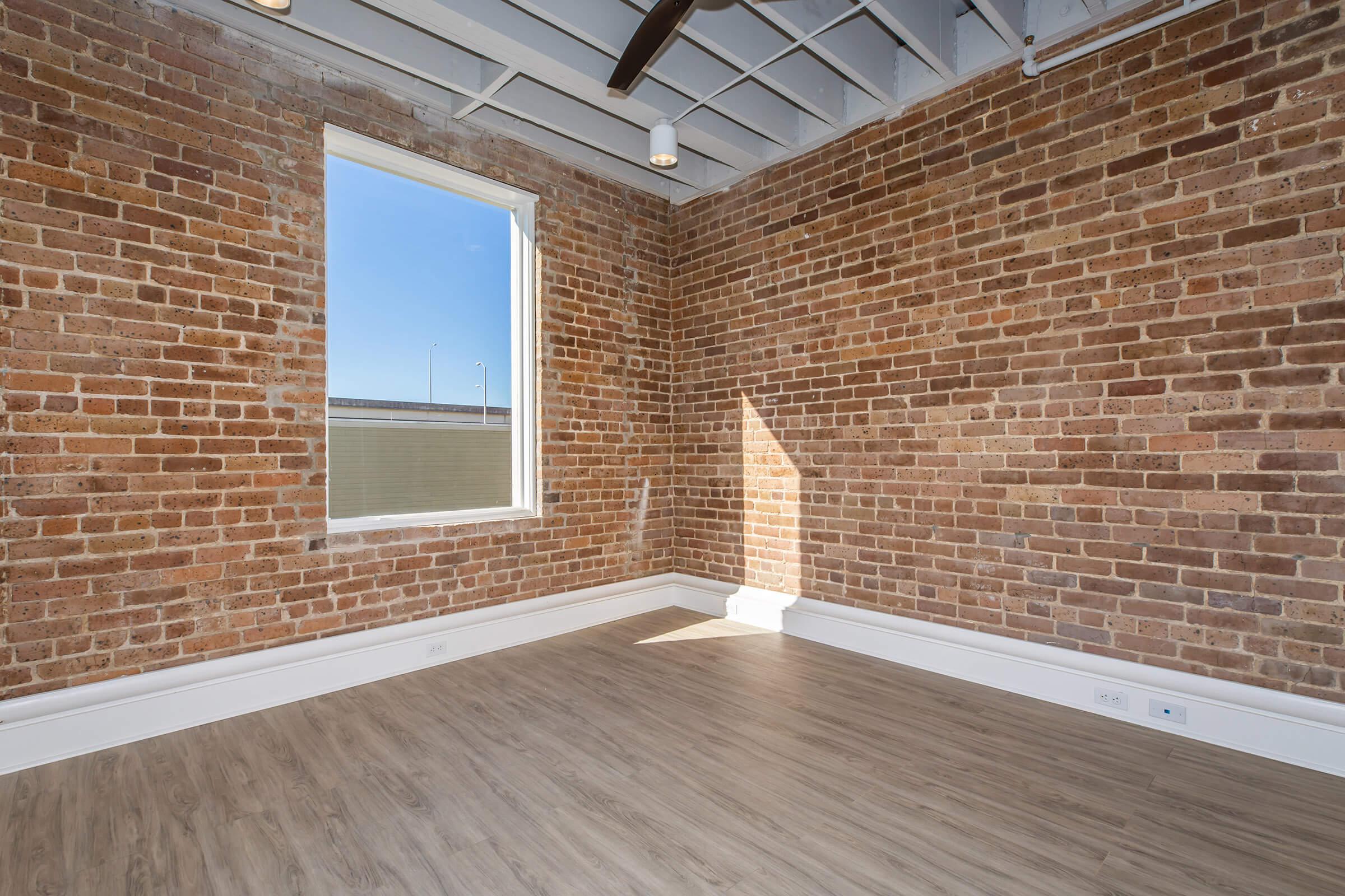 A spacious room featuring exposed red brick walls and natural light streaming in through a large window. The floor is a light wood finish, and the ceiling has a modern design with a ceiling fan. The overall ambiance is bright and inviting, ideal for various uses.