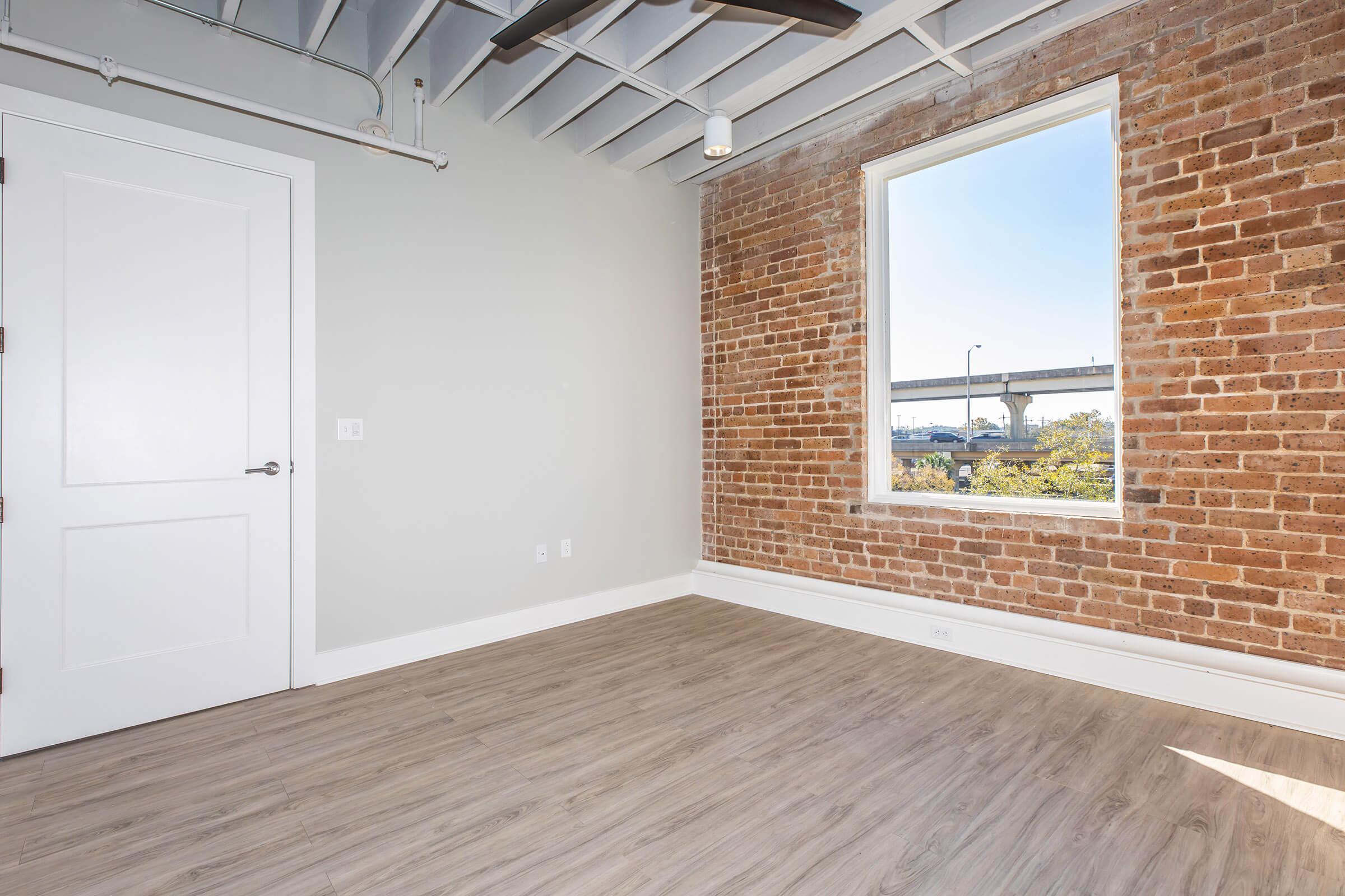 A modern room featuring a large window with a view of a bridge, exposed brick walls, and light wood flooring. The space is minimally furnished, with a white door and a ceiling fan visible. Sunlight illuminates the room, highlighting the blend of industrial and contemporary design elements.