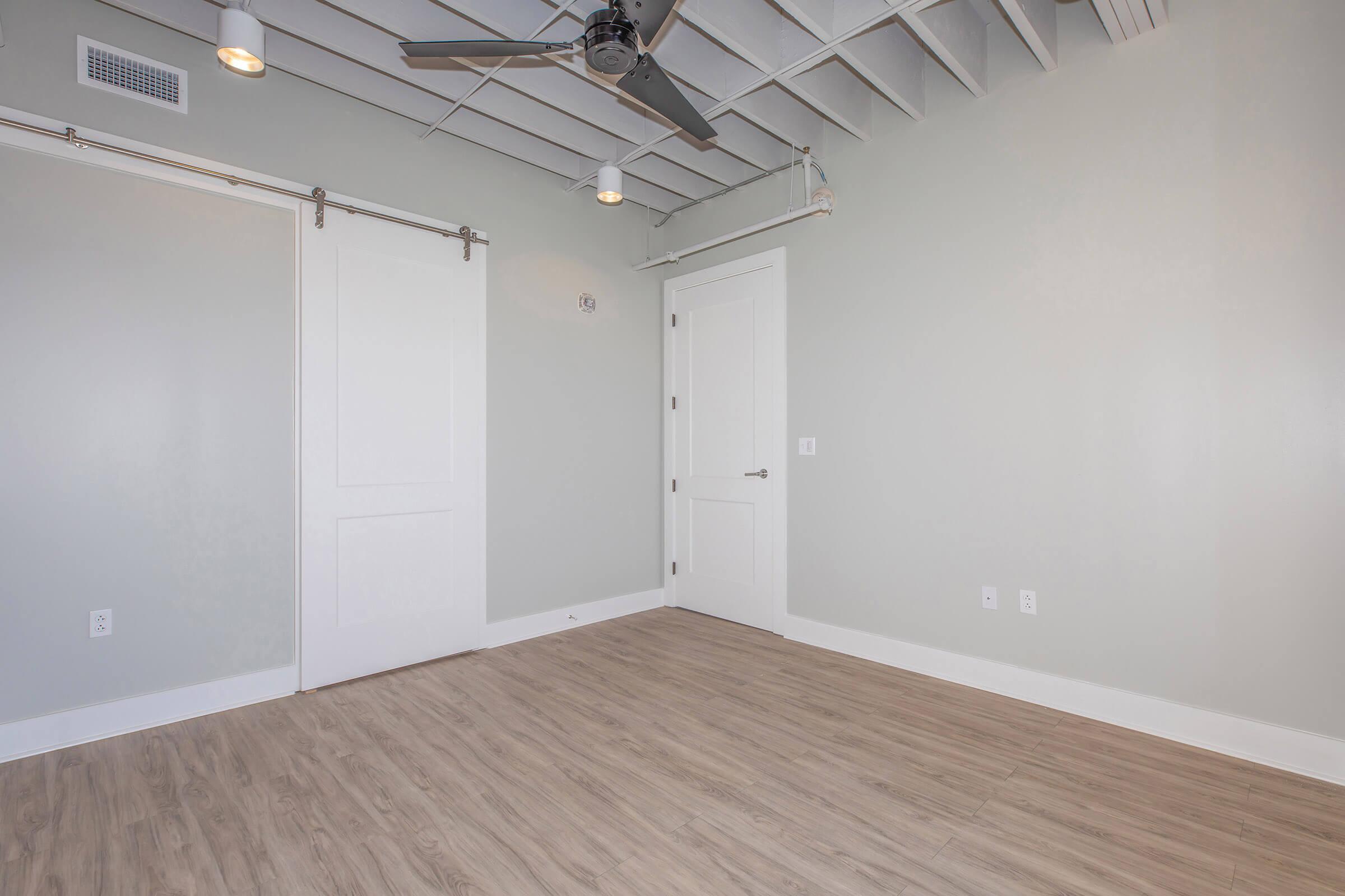 A spacious, empty room with light gray walls and wooden flooring. There are white sliding barn doors on the left and a closed white door on the right. A ceiling fan is mounted in the center, and overhead lights create a bright and airy atmosphere. The room has a modern and minimalistic design.