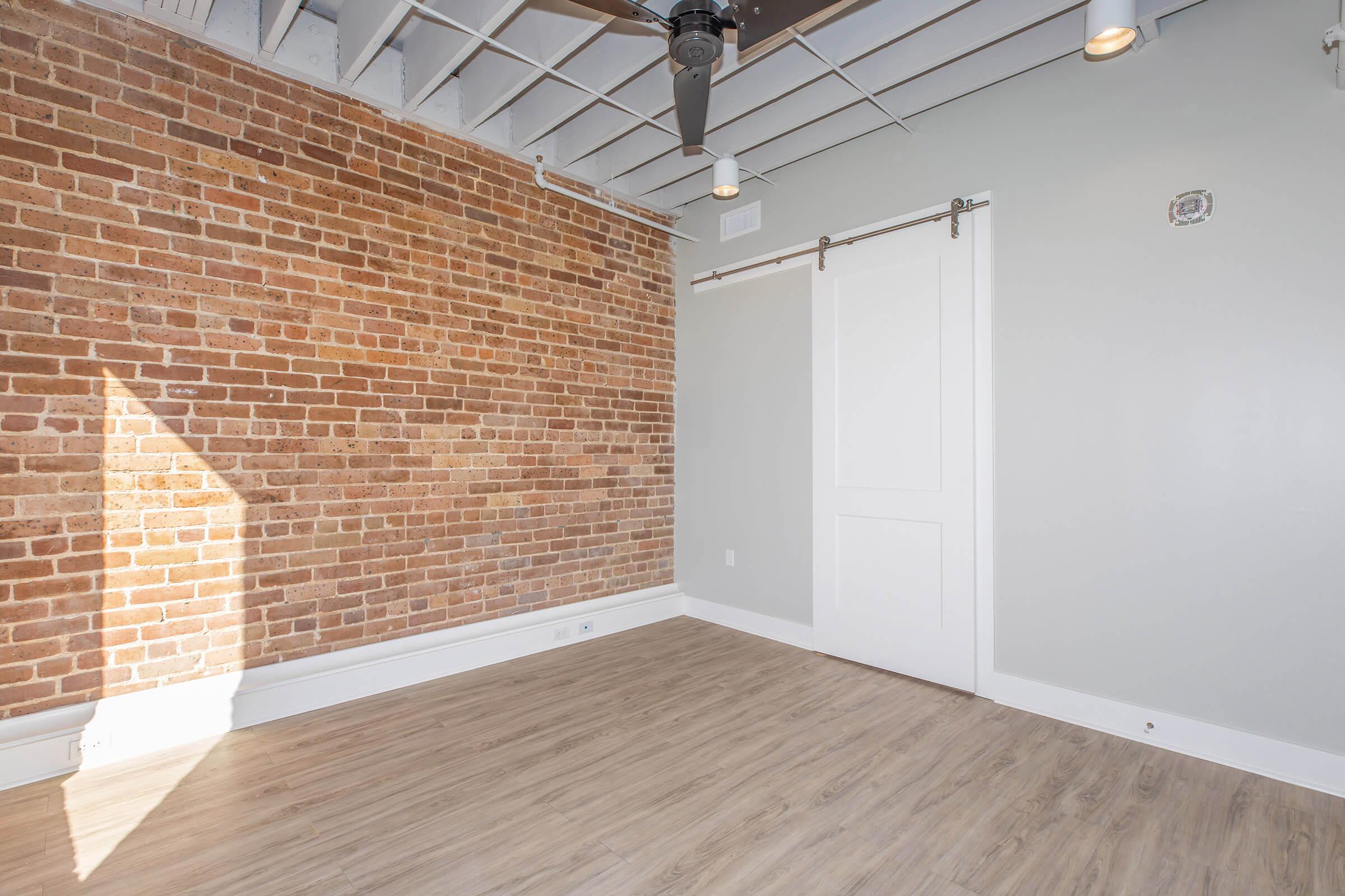 A spacious interior room featuring a rustic brick wall, light gray painted walls, and a sliding barn door. The ceiling is open with exposed beams and a ceiling fan. The floor is made of light-colored wood. Natural light enters from a large window, creating a bright and inviting atmosphere.