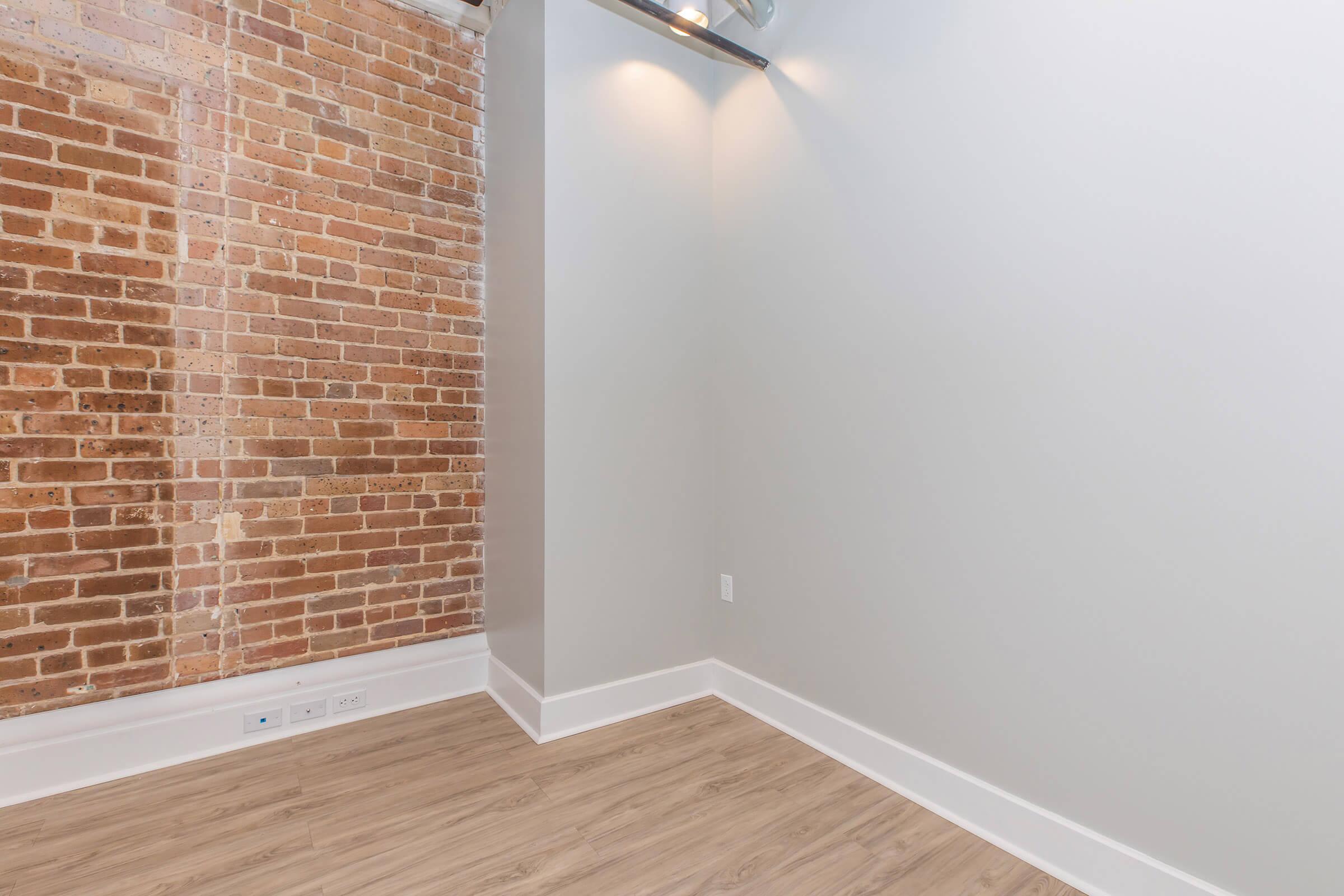 A spacious room featuring a textured brick wall on one side and a light-colored painted wall on the other. The floor is made of light wood, and there are two overhead lights illuminating the area. The space appears empty and minimalistic, suggesting versatility for various uses.