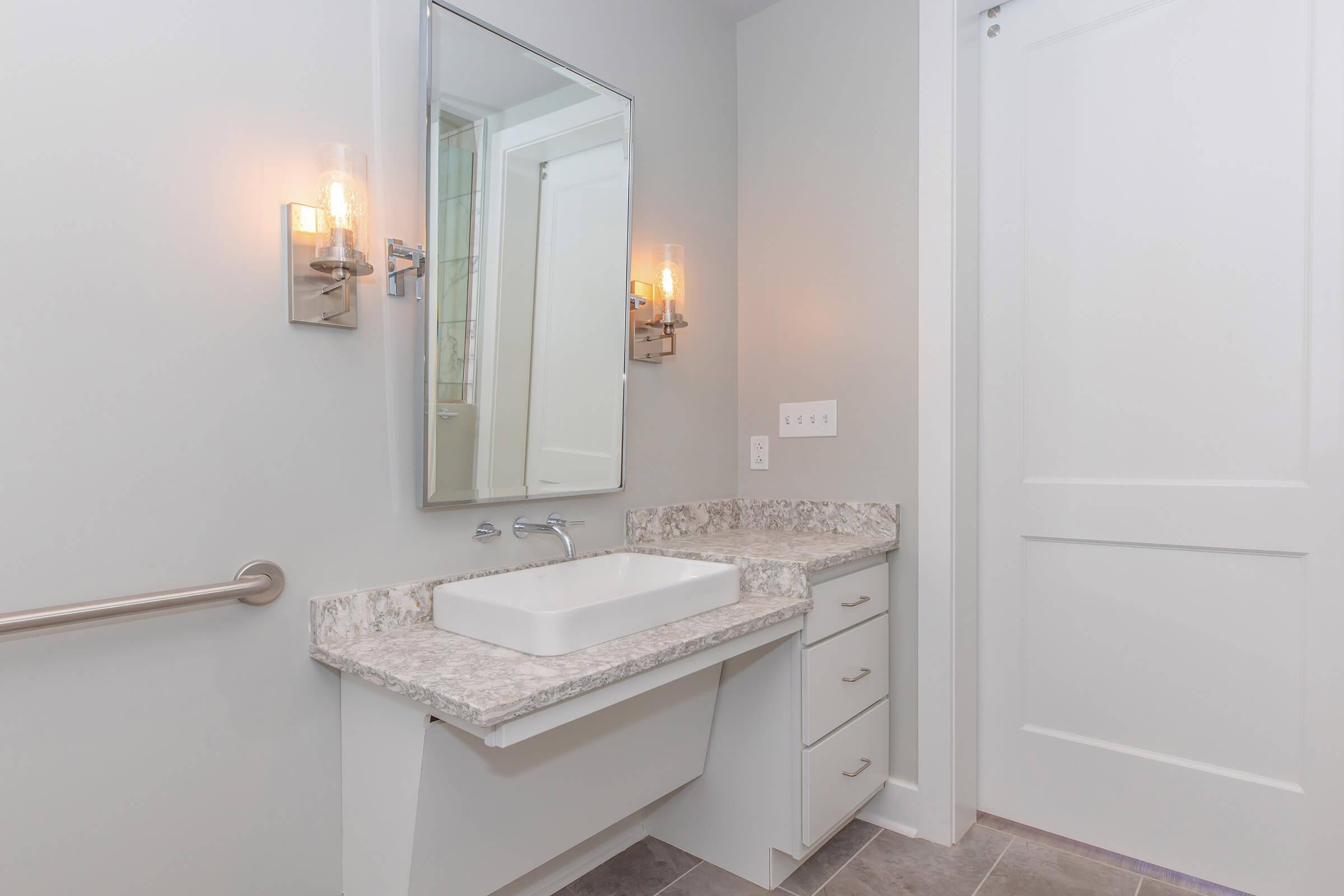 Modern bathroom featuring a sleek vanity with a rectangular sink, a wall-mounted mirror, and contemporary lighting fixtures. The color palette is light and neutral, with gray tile flooring and clean lines, creating a minimalist and spacious feel. A door can be seen partially closed in the background.