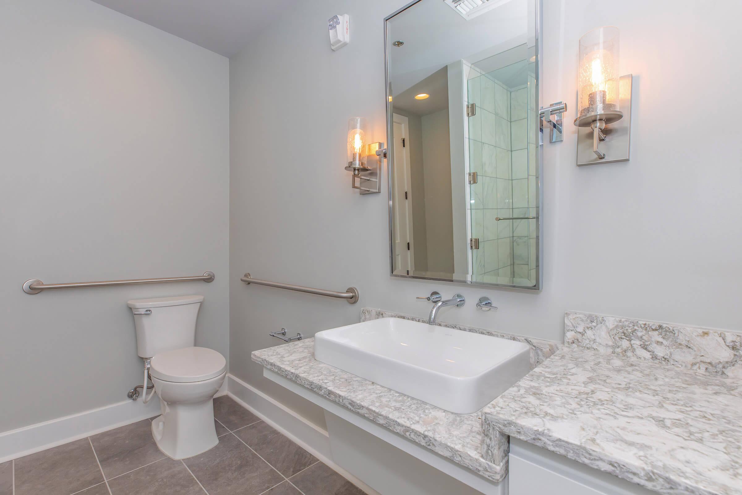 A modern bathroom featuring a white toilet, a sleek sink with a rectangular basin on a gray marble countertop, and a large mirror. The walls are painted gray, and there are stylish wall sconces providing light. Grab bars are installed for accessibility, and the floor is tiled in gray.