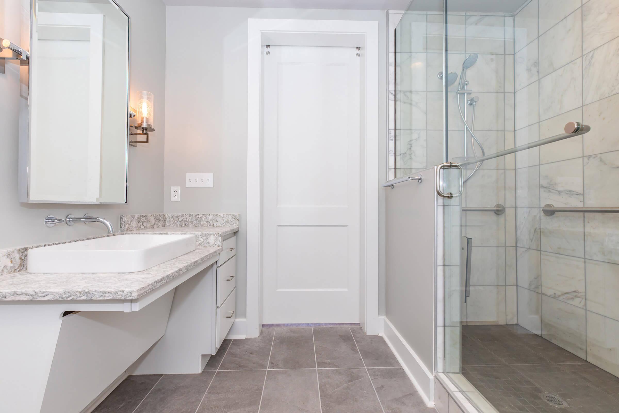 Modern bathroom featuring a double sink vanity with a granite countertop, a large mirror, and a walk-in glass shower with a marble tile design. The light gray walls and sleek fixtures contribute to a contemporary aesthetic. A white door leads to another area. The floor is covered in gray tiles.