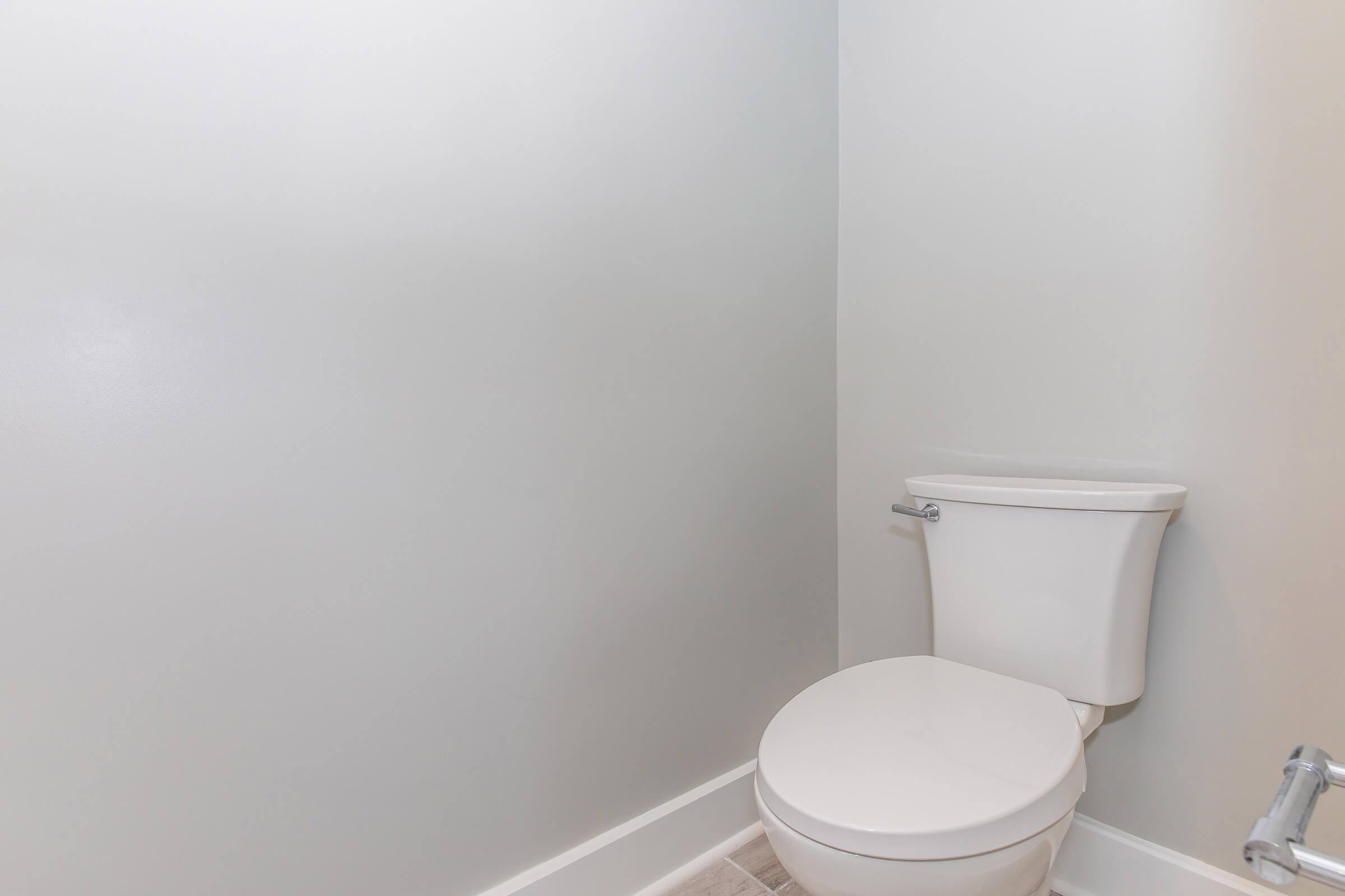 A clean, minimalist bathroom featuring a white toilet against a light gray wall. The floor is tiled in a neutral color, and the overall space is simple and uncluttered, emphasizing a modern design aesthetic.