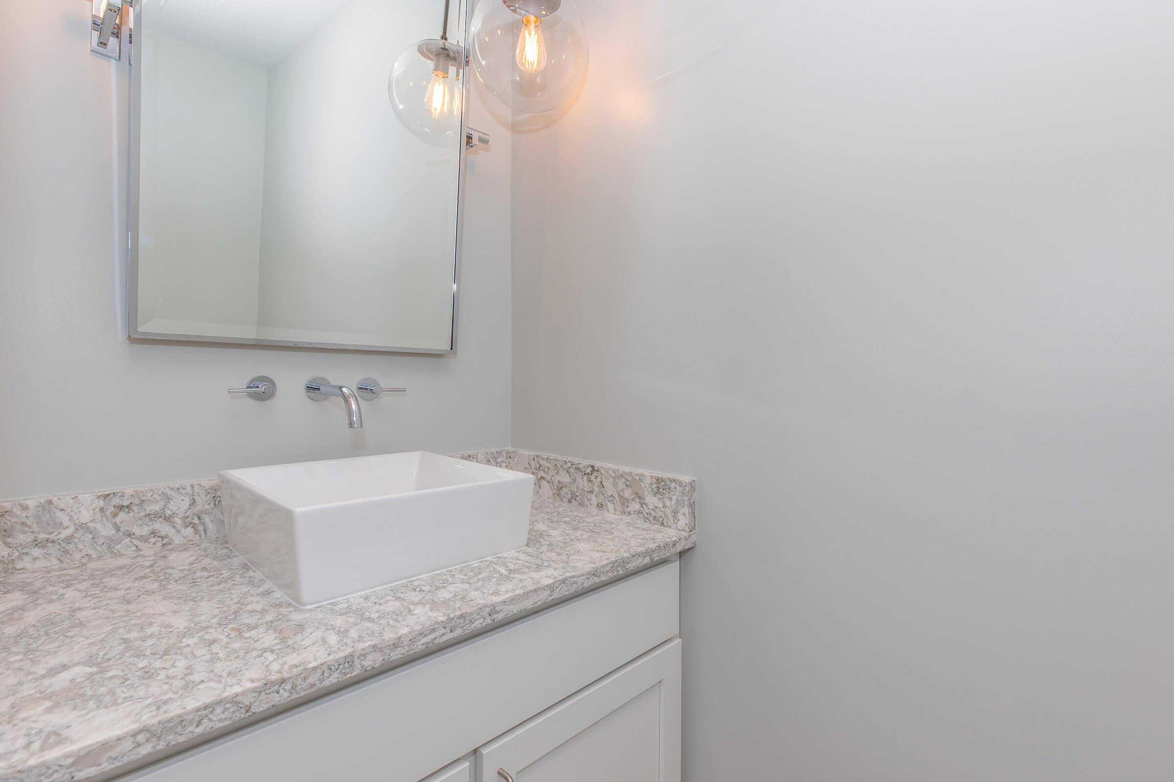 A modern bathroom with a white rectangular sink on a light-colored granite countertop. Above the sink, there is a large mirror flanked by an elegant light fixture. The walls are painted in a soft color, creating a clean and minimalist aesthetic.