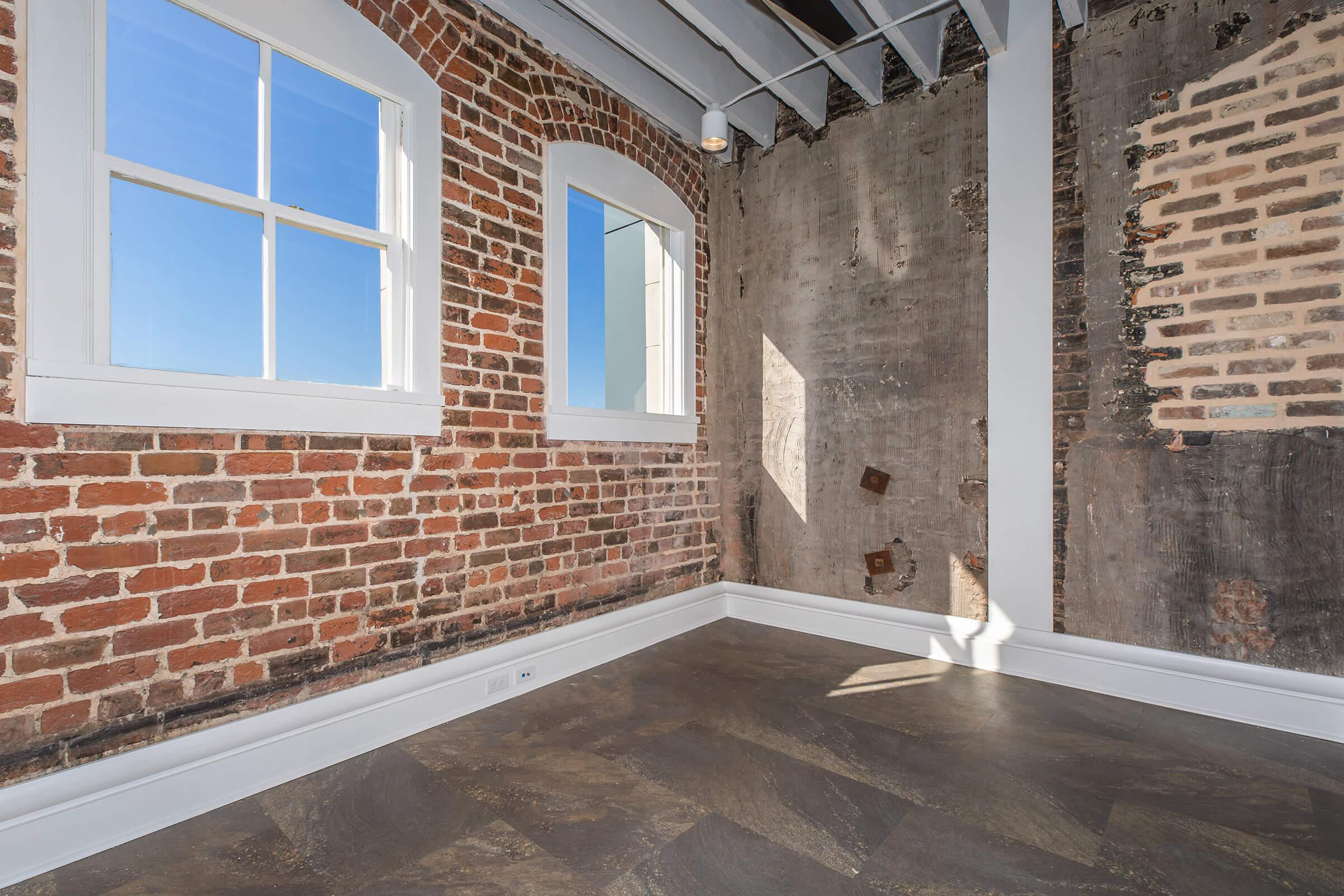 A bright, modern room featuring exposed brick walls and a polished floor. Large windows allow natural light to fill the space, highlighting the contrast between the rustic brick and the smooth, grayish wall. The ceiling has visible beams, adding to the industrial aesthetic.