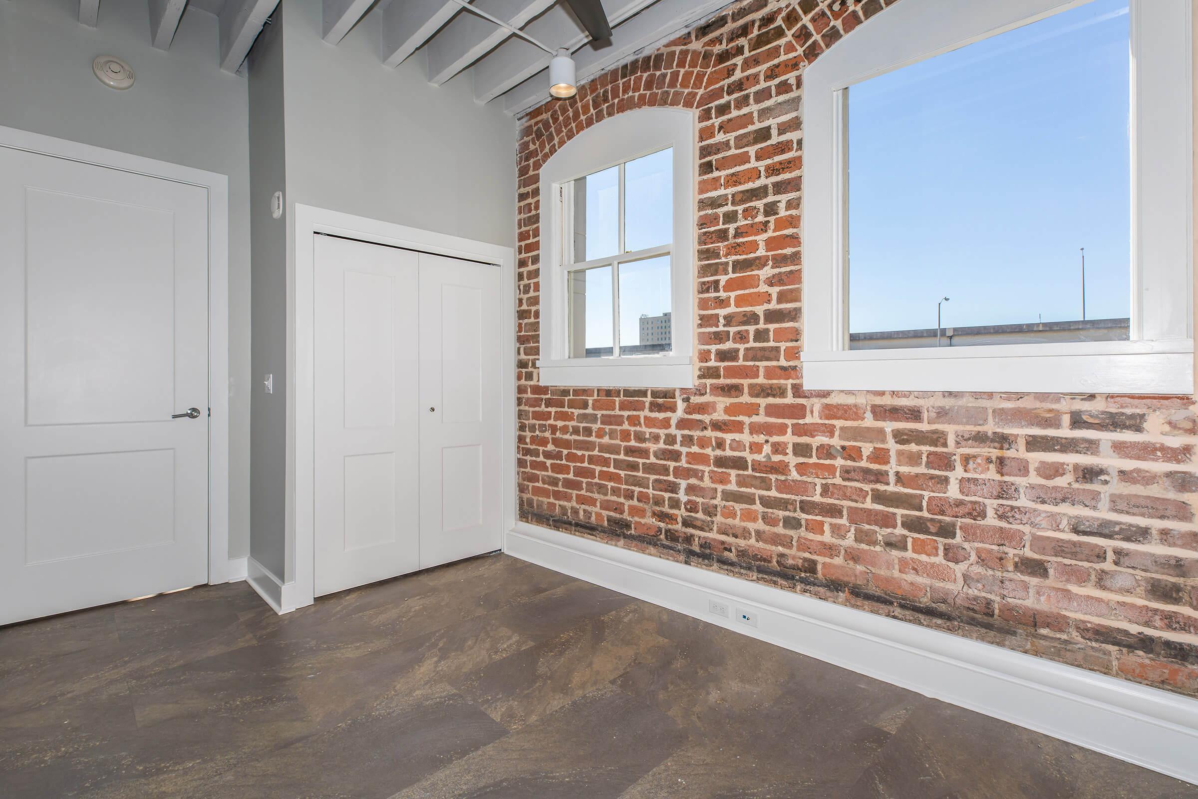 Interior view of a room featuring exposed brick walls, two large windows allowing natural light, a closet with double doors, and a modern gray floor. The ceiling has wooden beams, contributing to a contemporary yet rustic aesthetic.