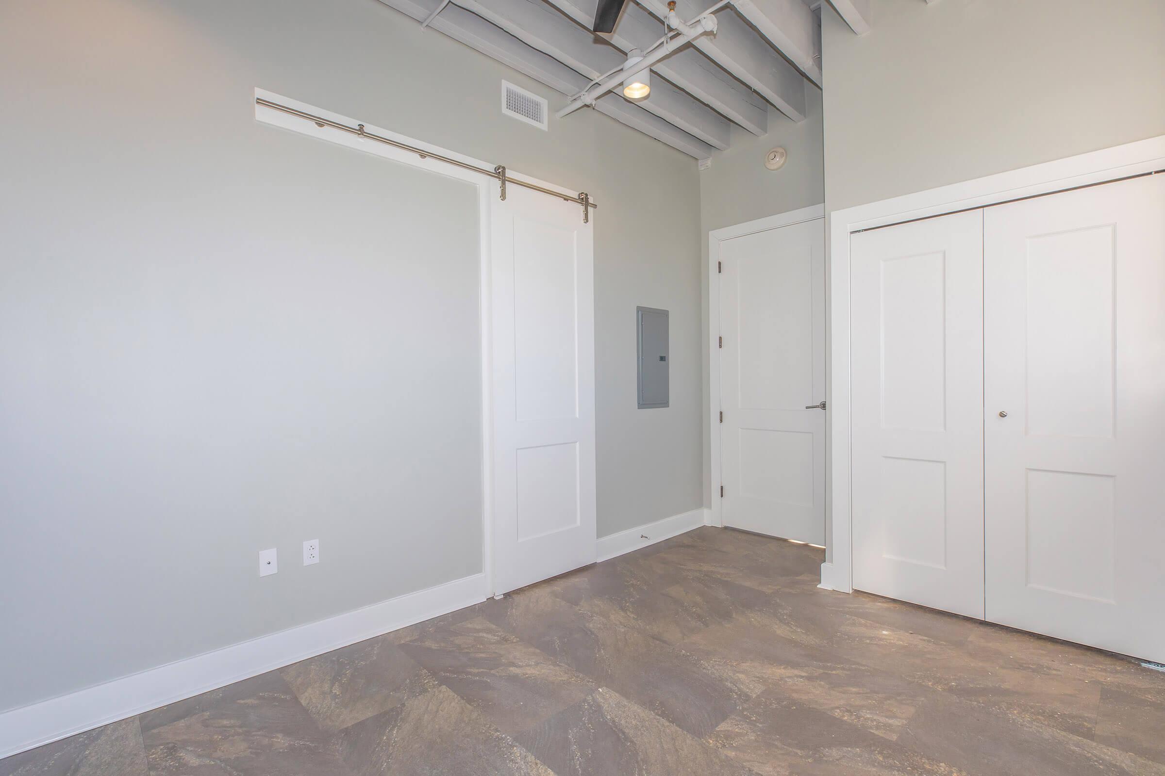 A spacious room with light gray walls and a high ceiling. It features a sliding barn-style door to the left, a closed closet with double doors on the right, and a gray stone floor. There is a mounted electrical panel on the wall, and natural light is coming in from an unseen window.