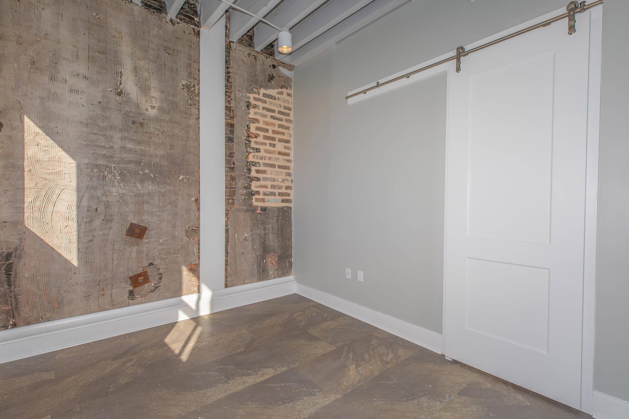 A spacious interior room with a combination of exposed brick and smooth gray walls. There's a sliding white door on one side and a large window casting natural light onto the floor, which features patterned tile. The ceiling is high with visible beams, adding a modern touch to the rustic elements.