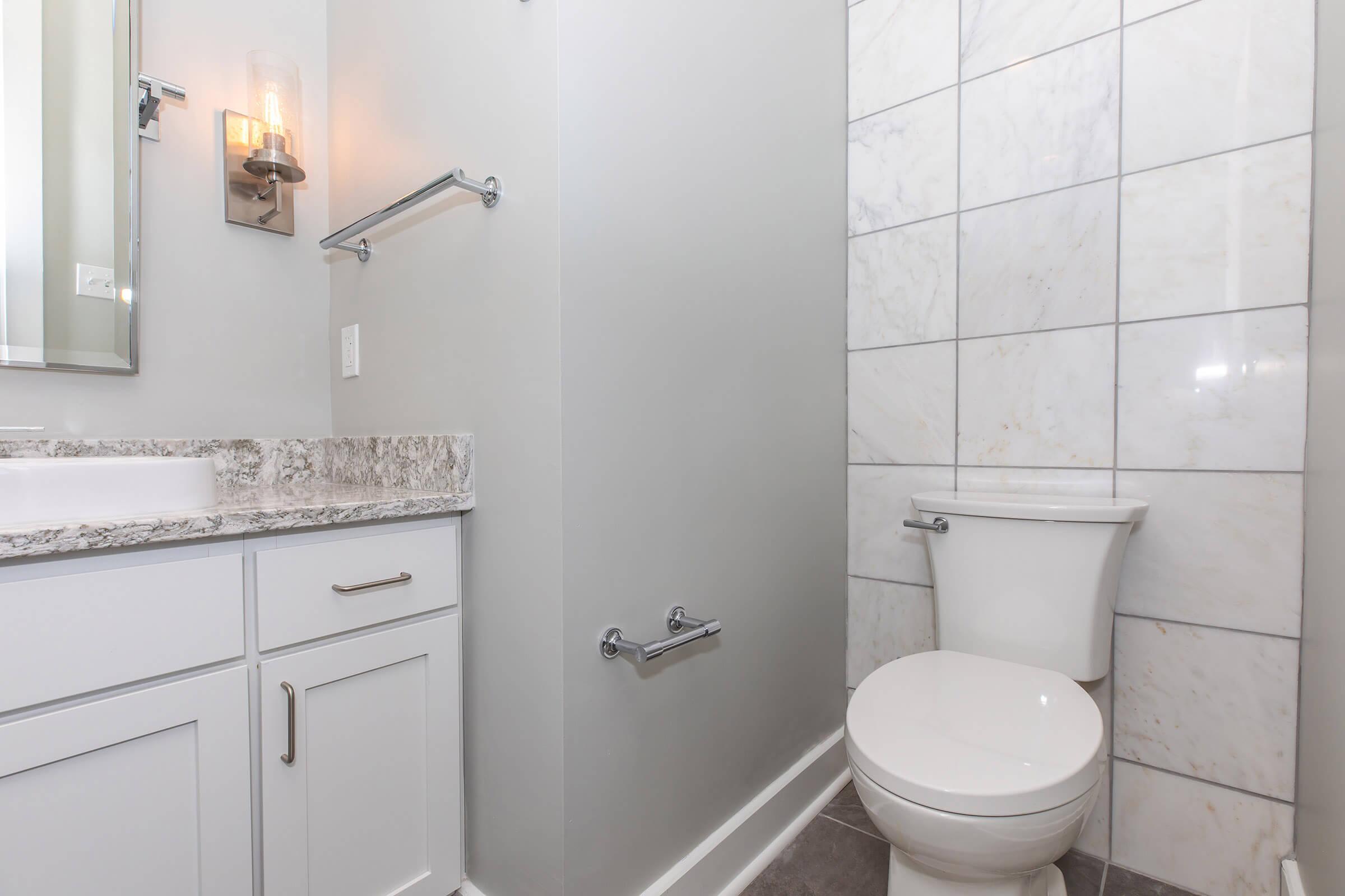 A modern bathroom with light gray walls, a white toilet, and a granite countertop sink. The wall features white tiles with a marbled design. A wall-mounted sconce is installed next to a mirror above the sink, and a towel rack is visible on the wall. The room is well-lit and has a clean, contemporary appearance.