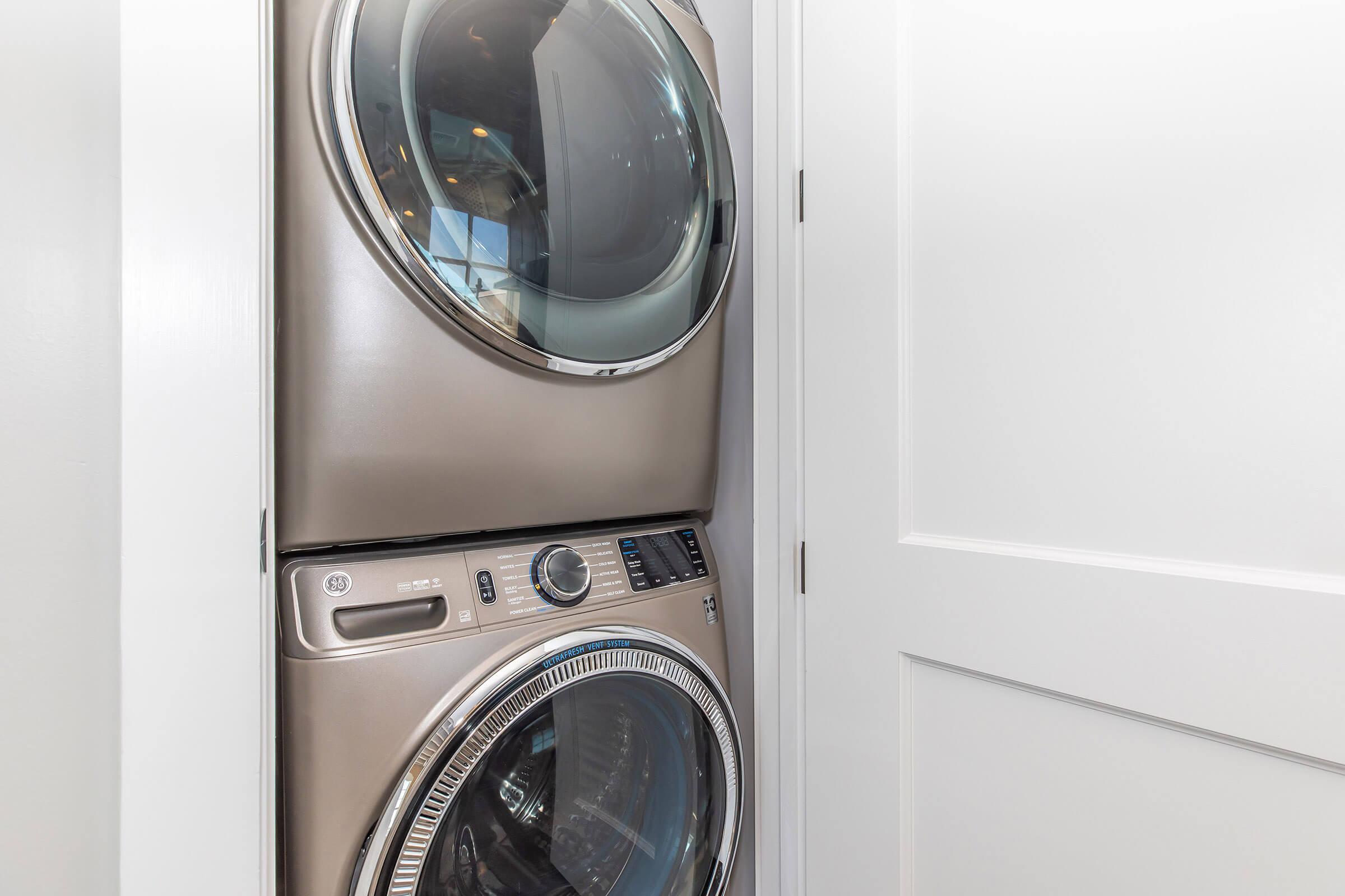 A modern stacked washer and dryer set in a small laundry space, placed against a white wall. The appliances have a sleek, metallic finish and are designed to save space in a home environment. A partially closed door is visible in the background.