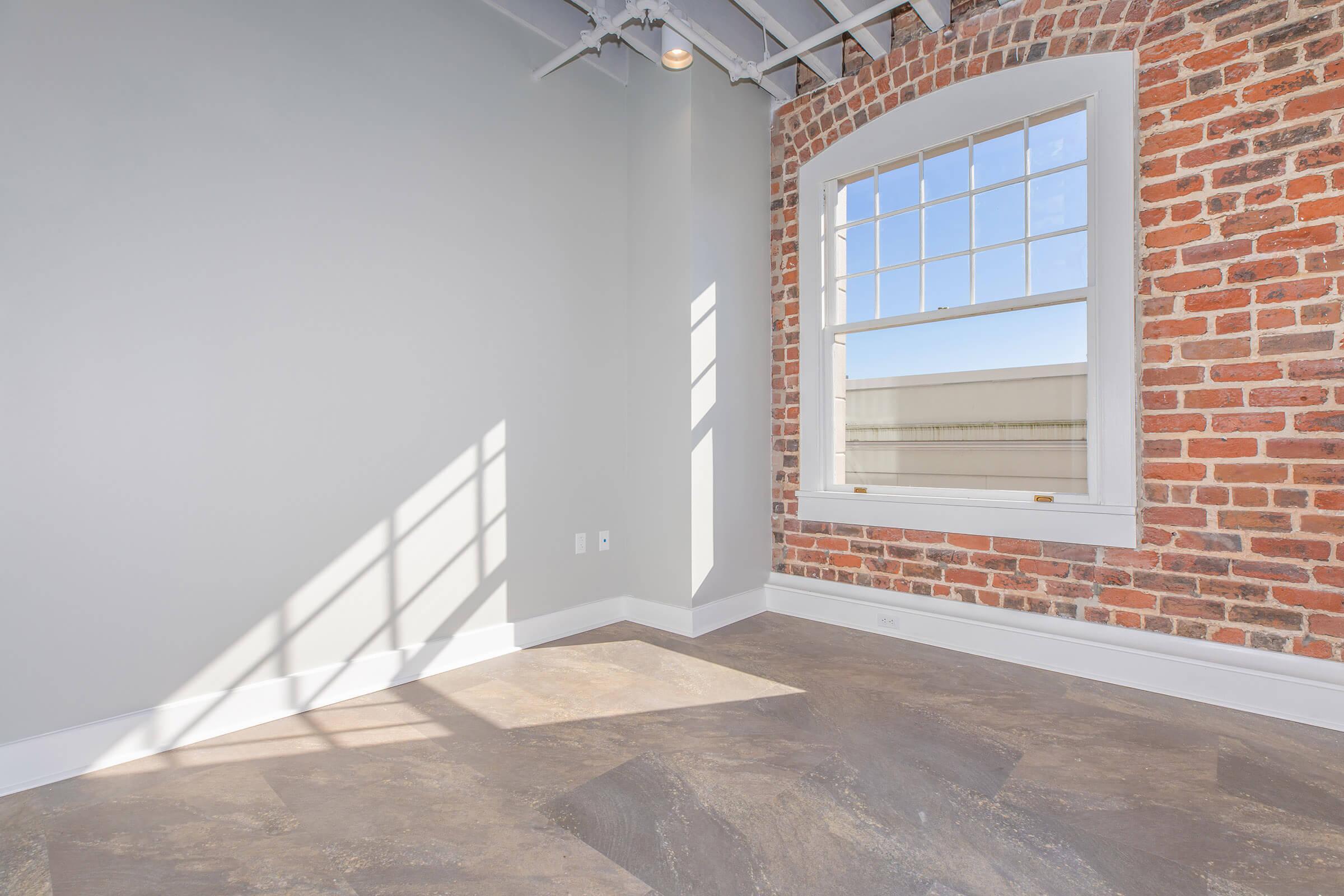 Bright and spacious room featuring a large window with natural light casting shadows on the floor. The walls are a mix of smooth light gray and exposed red brick, adding a modern yet rustic feel. The flooring is gray tile, complementing the overall aesthetic of the space.