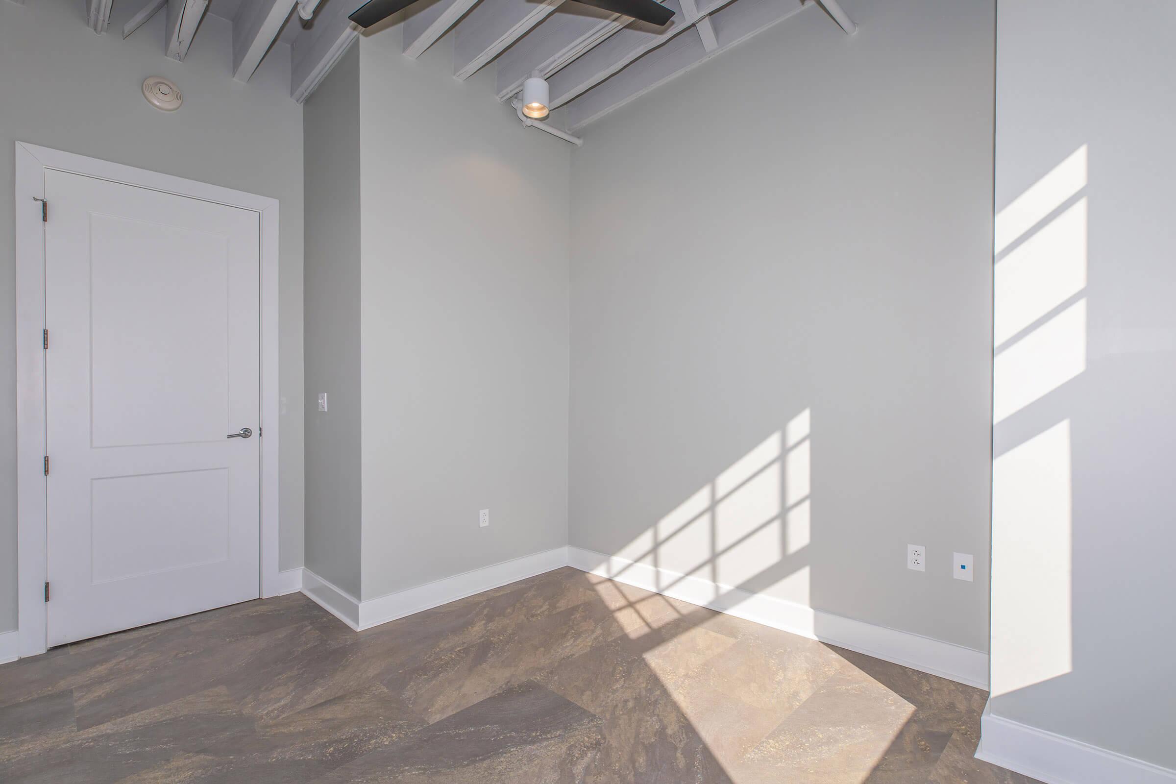 A well-lit, empty room with light gray walls and a modern ceiling featuring exposed beams. The floor has a textured design, and a door is visible on the left. Sunlight casts a shadow pattern on the floor from the window, creating a bright and airy atmosphere.