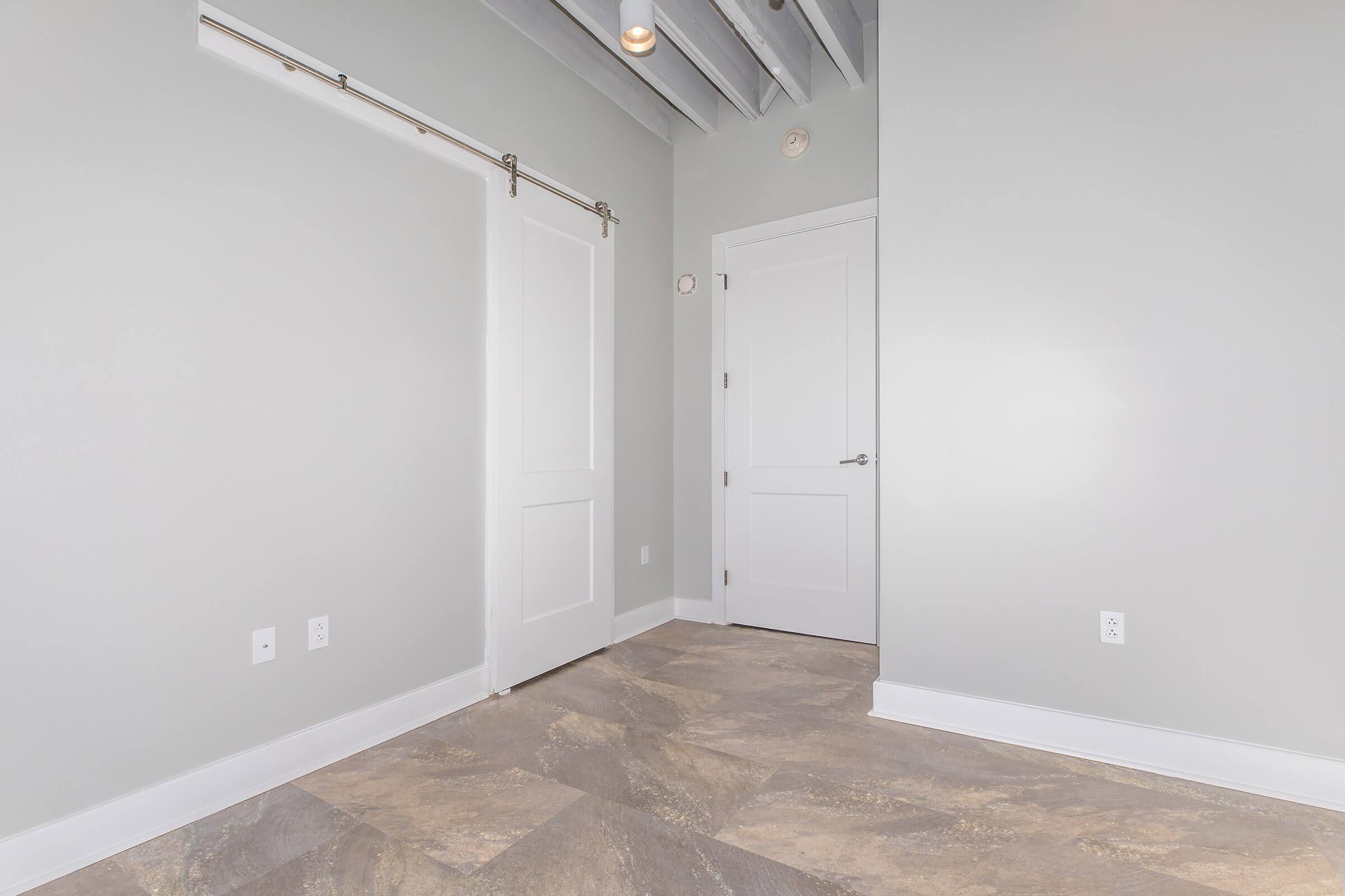 A well-lit, empty room featuring light gray walls, a textured floor, and exposed ceiling beams. One wall has a sliding door, while another wall includes a standard door. The space is clean and modern, suitable for various uses.