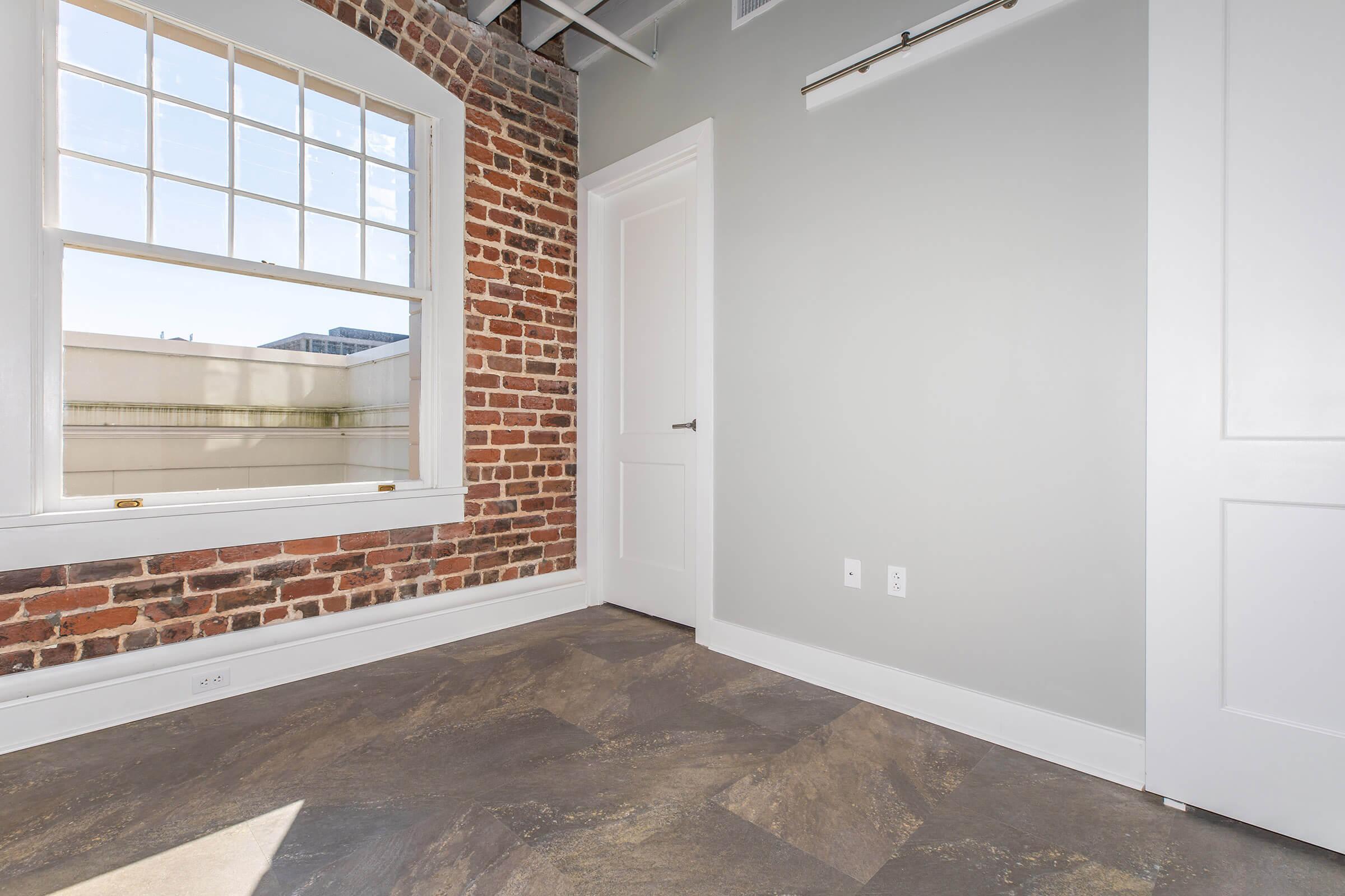 Interior of a room featuring a brick accent wall, large window allowing natural light, and a door. The floor is tiled with a stone-like pattern, and the walls are painted light gray. The ceiling is exposed, adding a modern touch to the space.