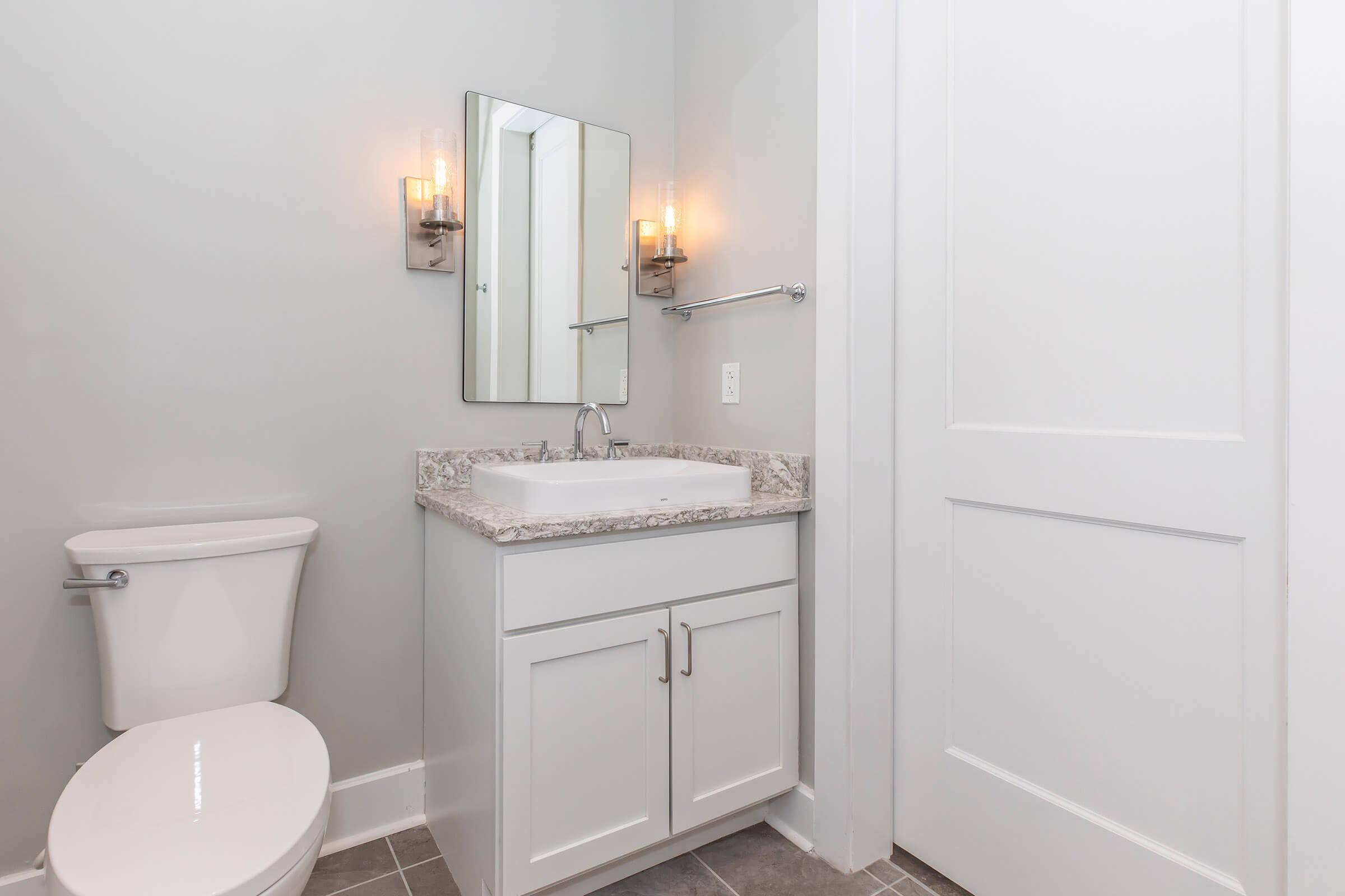 A modern bathroom featuring a white toilet, a single sink vanity with granite countertop and white cabinetry, a large mirror above the sink, and two wall sconces providing soft lighting. The walls are painted light gray, and a door is visible on the right side. The floor is tiled in a neutral color.