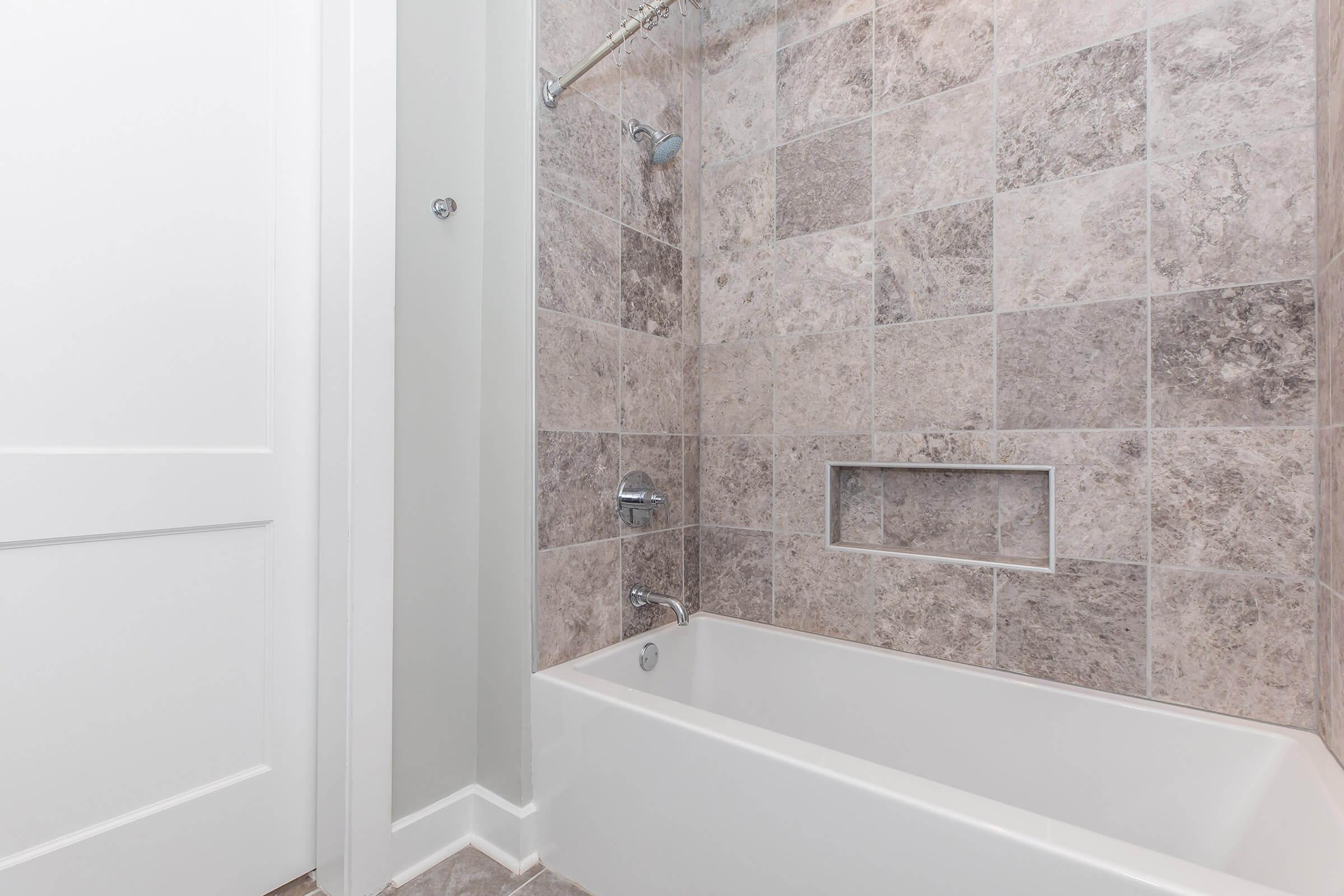 A clean bathroom featuring a modern bathtub and shower area. The walls are covered with large gray and beige stone tiles. A sleek showerhead and faucet are visible, along with a built-in niche for toiletries. The room has a bright and airy feel, complemented by a white door and trim.