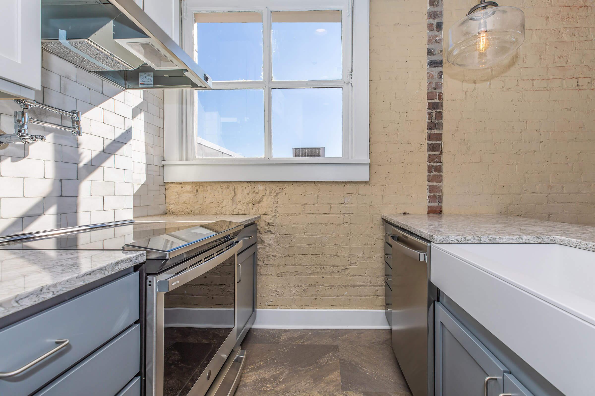 Modern kitchen featuring light gray cabinets, a stone countertop, stainless steel appliances, and a spacious window. The wall has exposed brick, adding a rustic touch, while natural light streams in, creating a bright and inviting atmosphere.