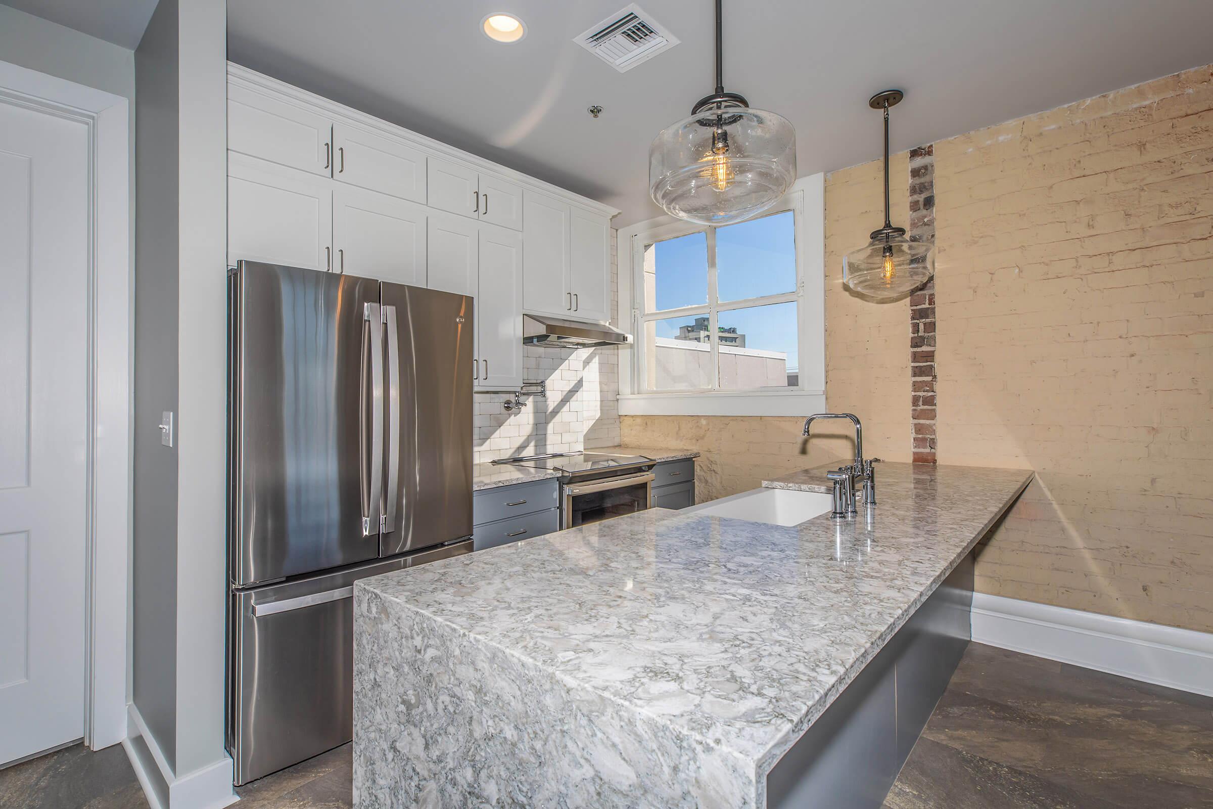 A modern kitchen featuring a large island with a grey marble countertop, stainless steel appliances, including a refrigerator and oven, white cabinetry, and exposed brick walls. Large windows allow natural light to fill the space, enhancing the contemporary design.