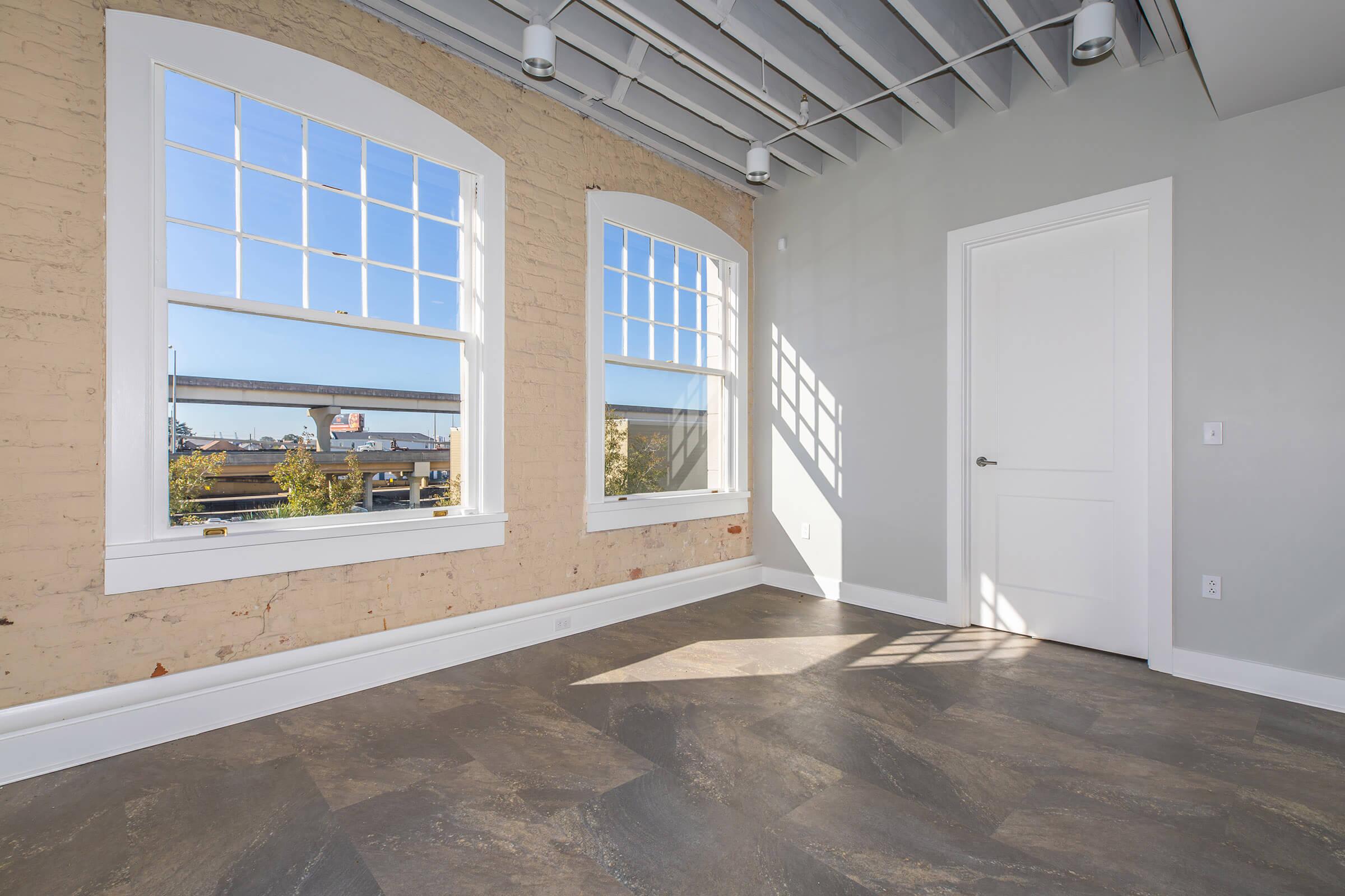 A bright, airy room featuring a light gray wall with large windows allowing natural light to fill the space. The floor has a gray stone-like pattern, and there’s a white door on the right. The walls are partially exposed brick, adding a touch of industrial charm.