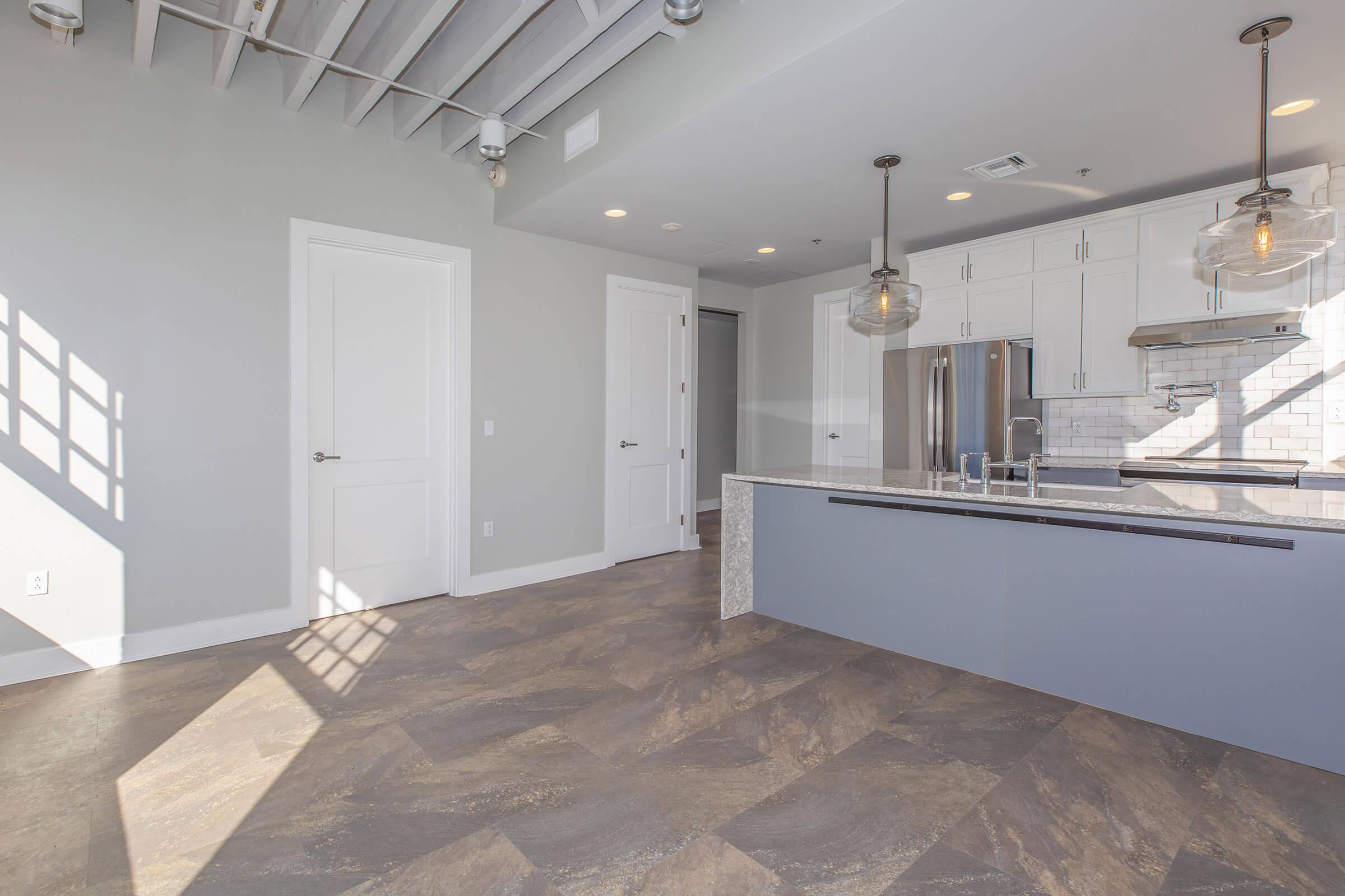 Modern kitchen with an island featuring a gray-blue base. The space has light-colored cabinets, stainless steel appliances, and a tiled backsplash. Large windows allow natural light to fill the room, casting shadows on the floor. The floor is a mix of brown and gray tiles, and there are double doors leading to another room.