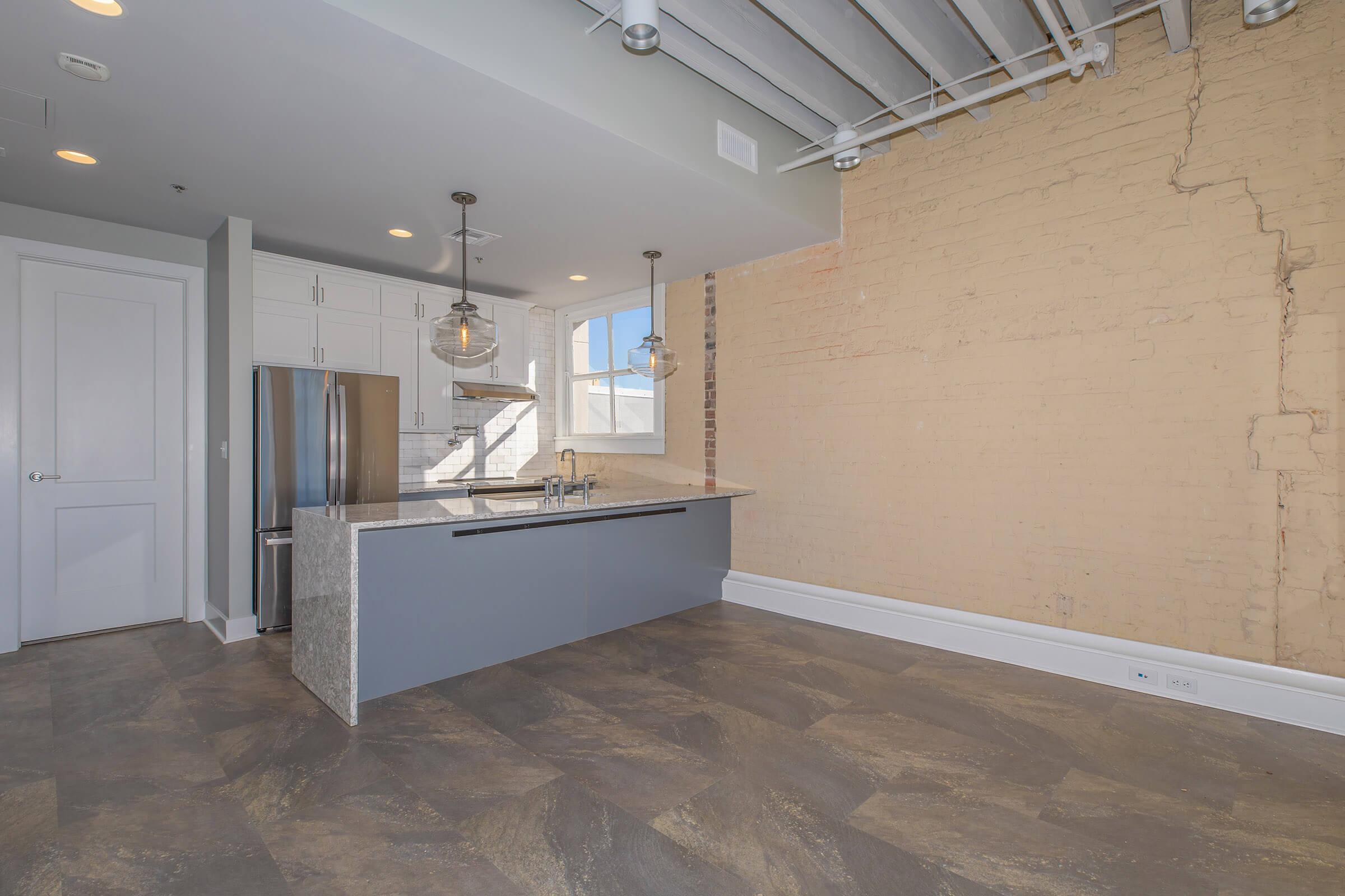 Modern kitchen with an island featuring a gray countertop and silver fixtures. There are white cabinets, a stainless steel refrigerator, and industrial-style pendant lights hanging from the ceiling. The wall is exposed brick, adding a rustic touch, while large windows provide natural light. The flooring is dark and textured.