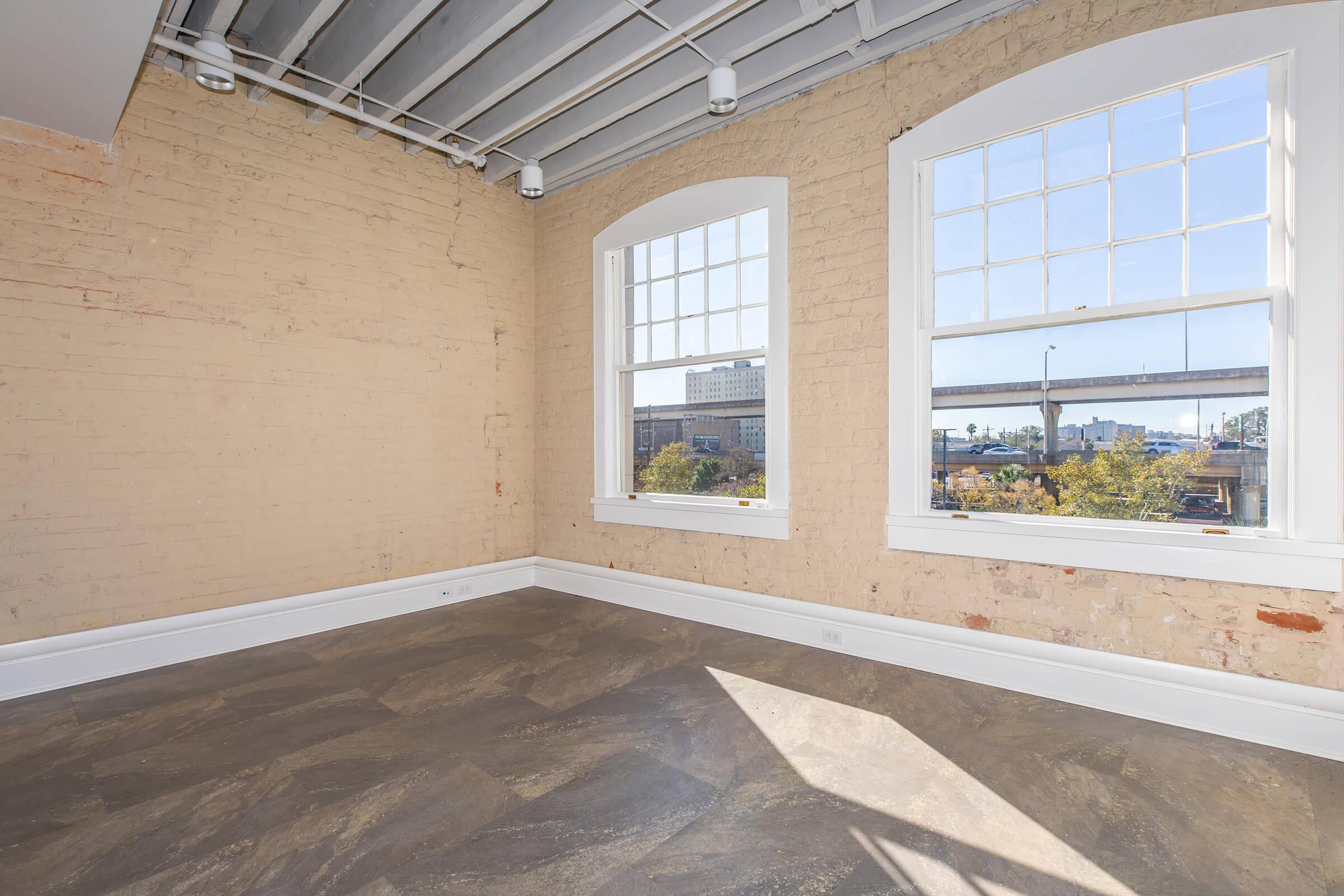 A bright, empty room with exposed brick walls and large windows letting in natural light. The floor is a modern, polished stone surface. The view outside the windows shows a cityscape with a bridge in the background, indicating an urban setting. The ceiling features industrial-style beams.