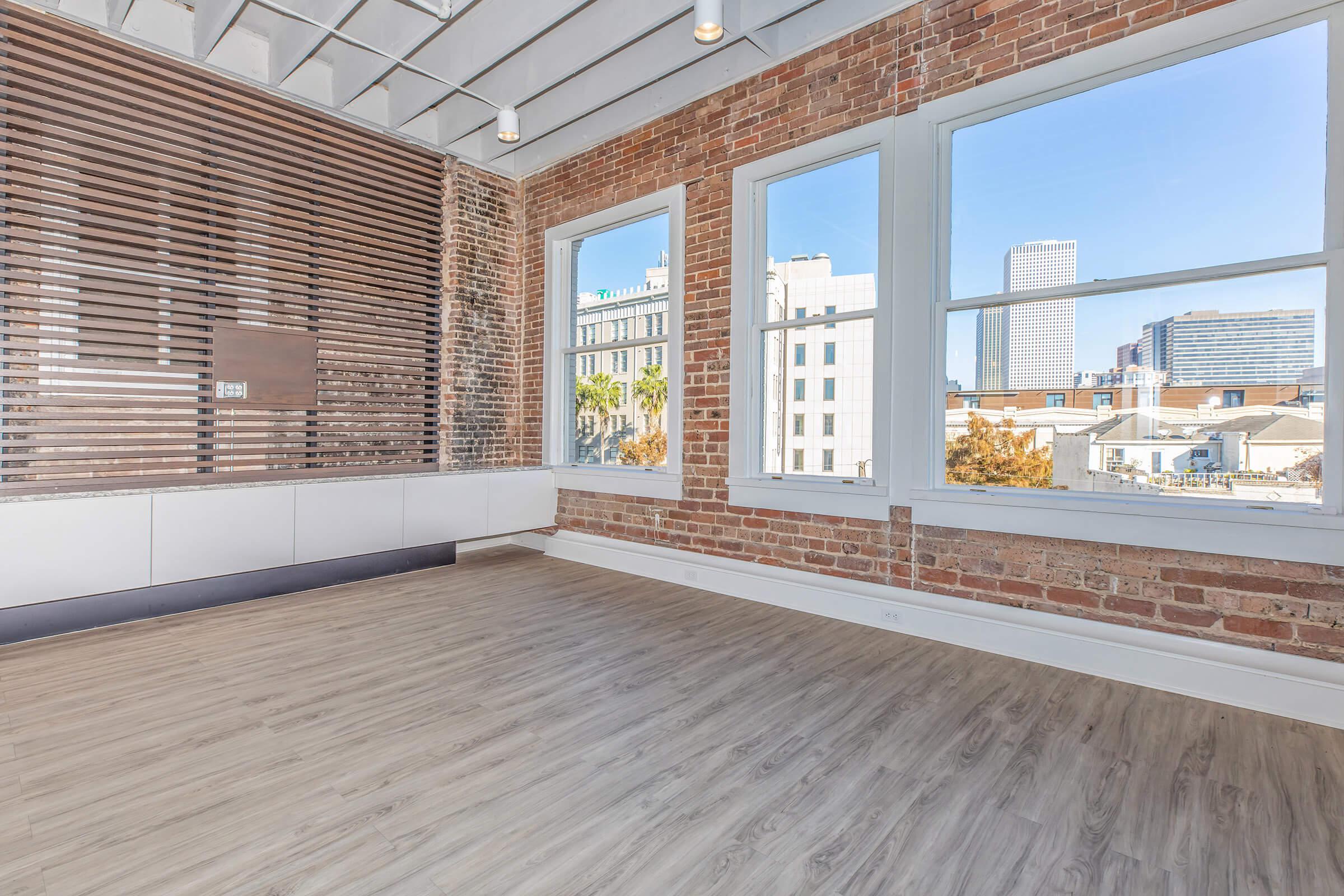 A spacious room featuring exposed brick walls, large windows allowing natural light, and a modern wooden floor. The view outside showcases a city skyline with tall buildings and blue skies. There is a decorative wooden wall element on one side, adding to the contemporary aesthetic.