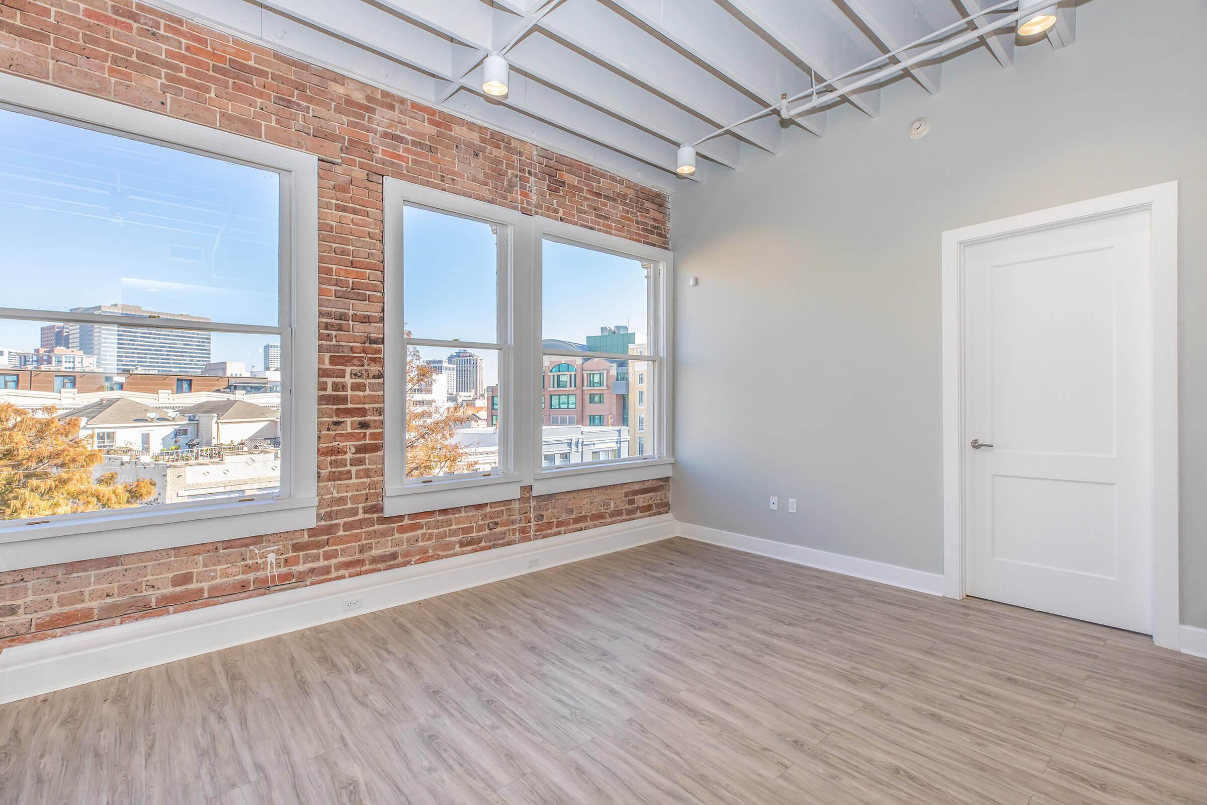 A spacious, modern room with large windows offering a view of a city skyline. The walls feature exposed brick, and the ceiling has metallic beams. The floor is light-colored wood, creating a bright and airy atmosphere. A white door is visible on one side of the room.