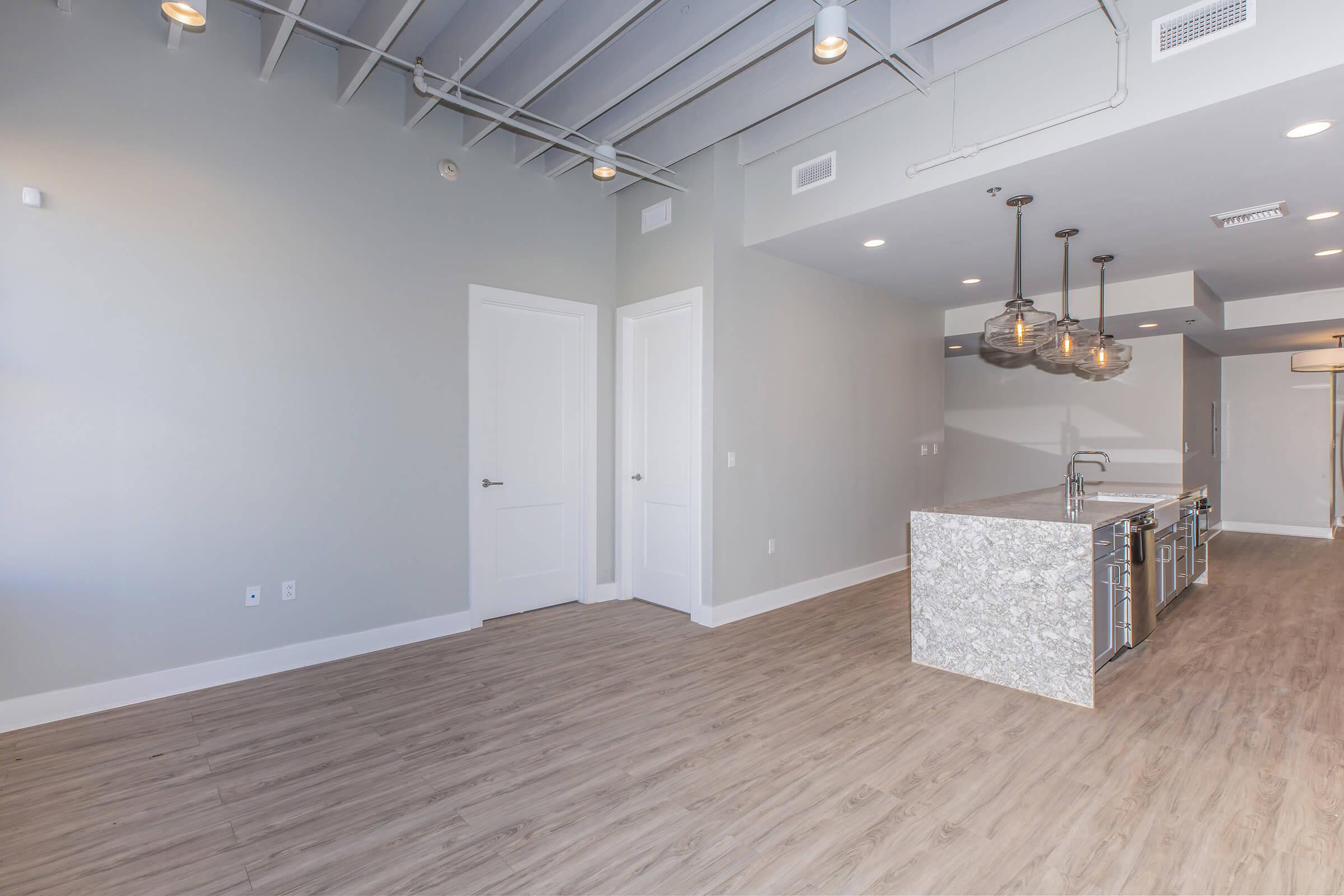 A modern, spacious living area with light-colored walls and wood-like flooring. To the left, there’s a doorway leading to another room. The kitchen features a large island with a countertop made of light-colored stone, and stylish pendant lights hang above. The overall design is open and contemporary.