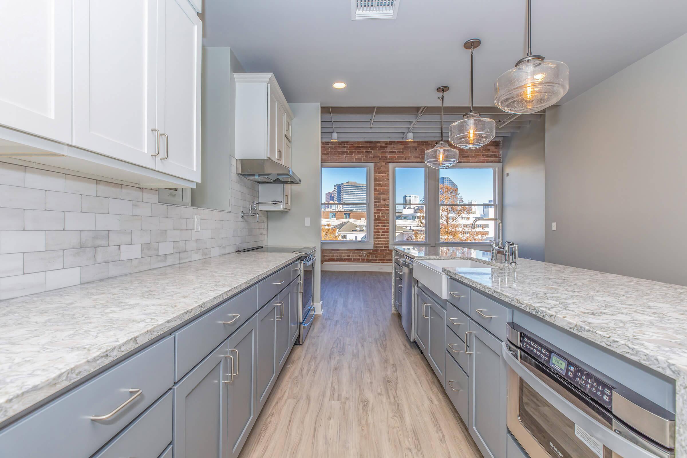 Modern kitchen featuring white cabinets and gray lower cabinets with a marble countertop. There are stainless steel appliances, including a built-in oven and a dishwasher. Large windows offer a view of a cityscape, and the room has pendant lighting hanging from the ceiling.