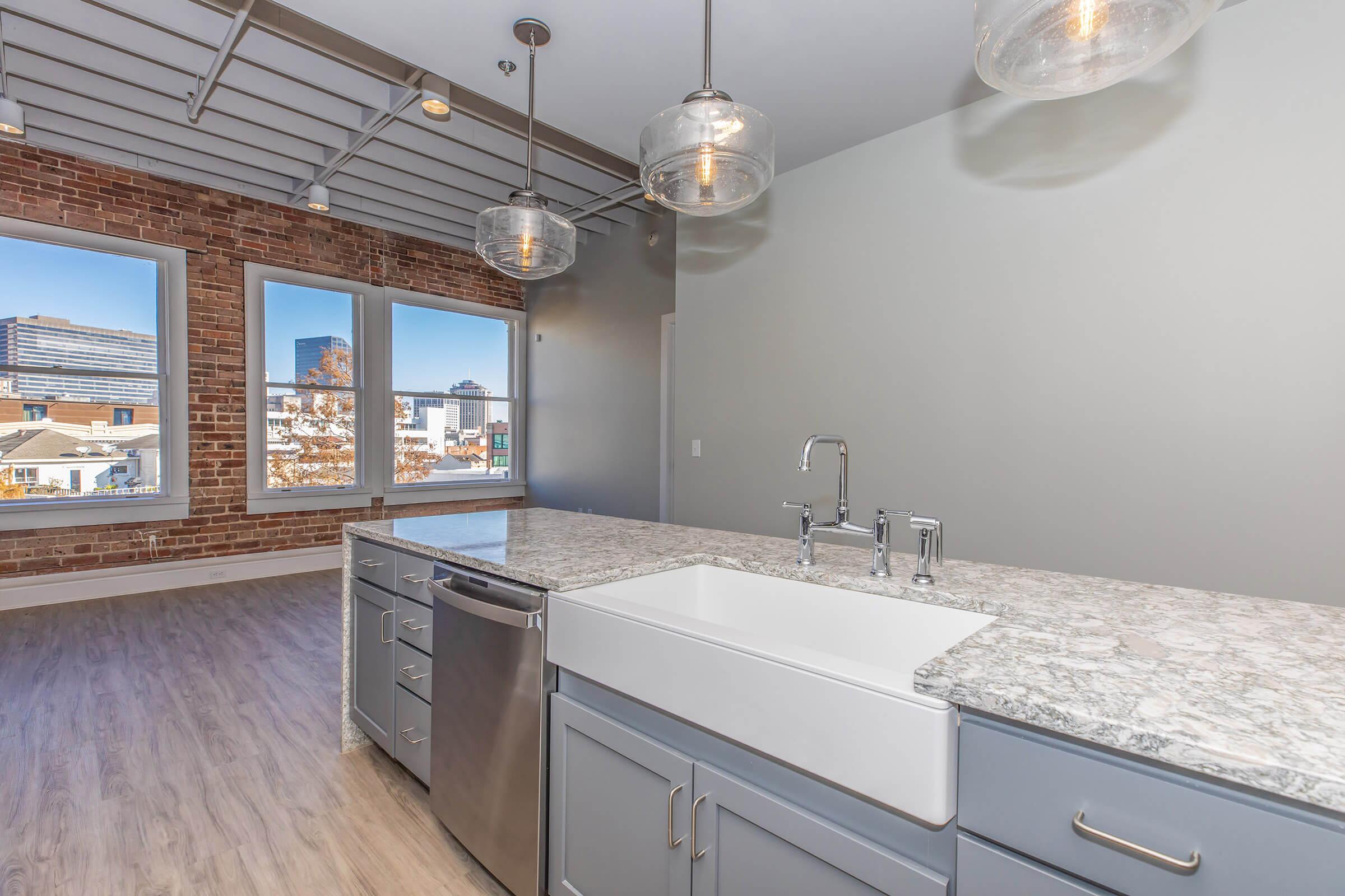 A modern kitchen featuring a spacious granite countertop with a farmhouse sink and stainless steel dishwasher. The background includes exposed brick walls and large windows showcasing a cityscape. Pendant lighting hangs from the ceiling, adding a contemporary touch to the open space.