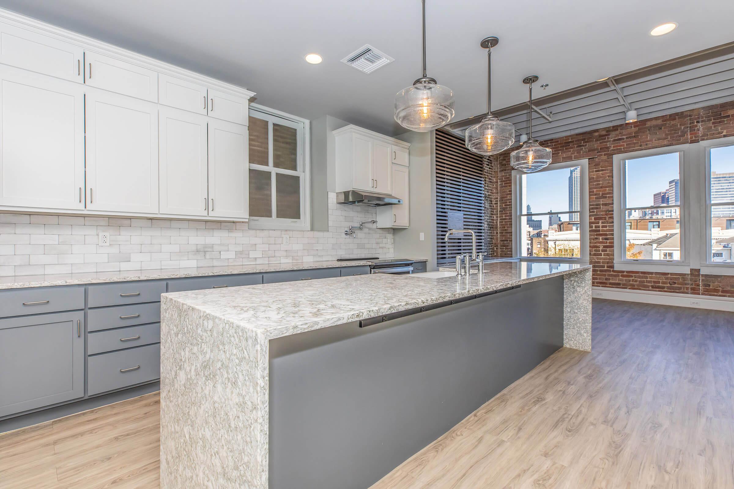 Modern kitchen featuring an island with a gray countertop, sleek cabinetry in white and gray, and a backsplash of white tiles. The space has large windows offering a view of the city and exposed brick wall accents. Stylish pendant lights hang above the island, enhancing the contemporary design.