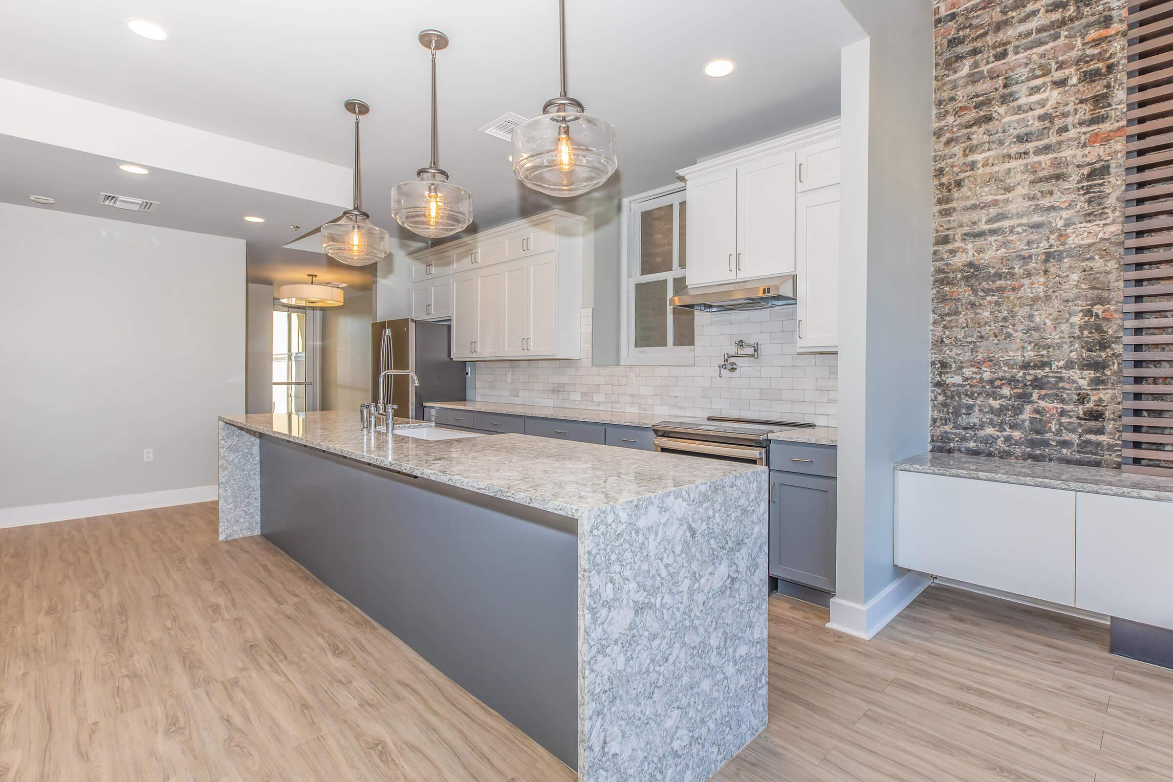 A modern kitchen featuring a large island with a marble countertop, pendant lighting above, and a blend of white and gray cabinetry. The backsplash is tiled, and there are exposed brick accents on one wall, complementing the light wood flooring and spacious design.