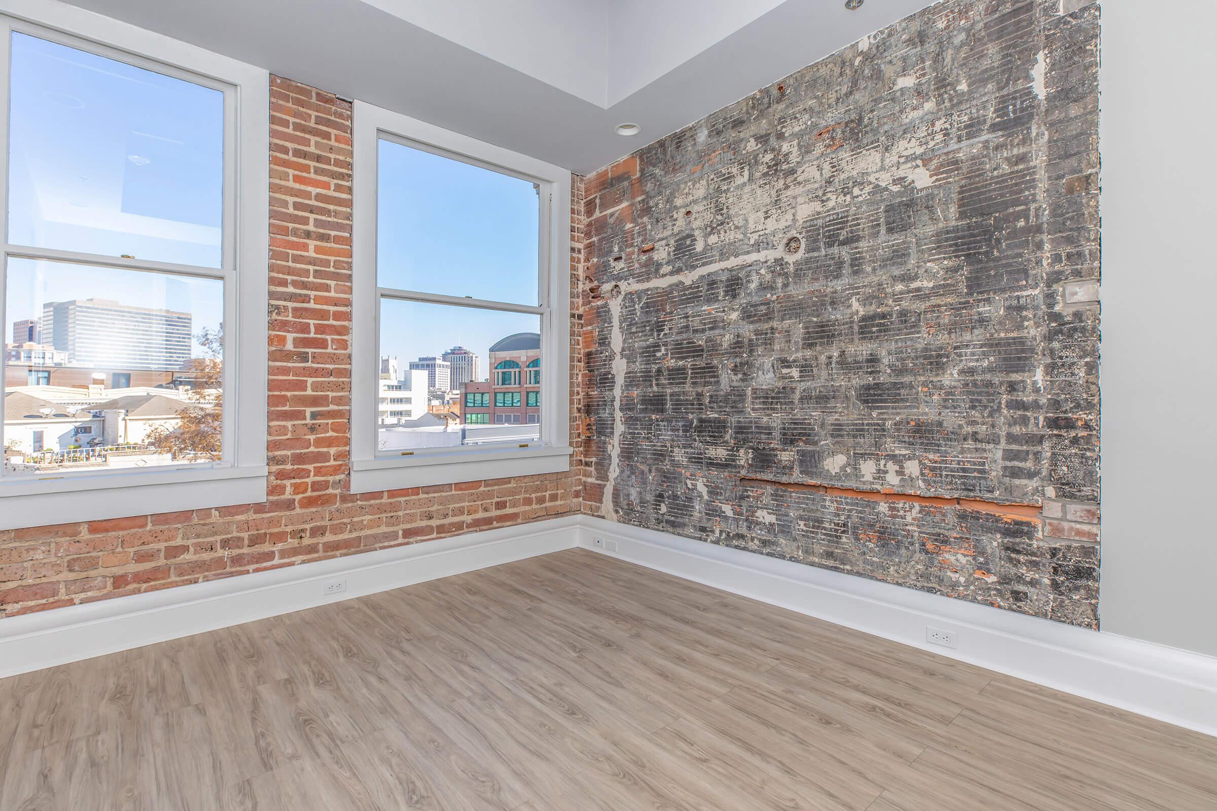 A well-lit room with two large windows showcasing a city view. The room features a combination of exposed brick walls and a textured gray wall, with wooden flooring. The space appears modern and empty, ready for furnishing.