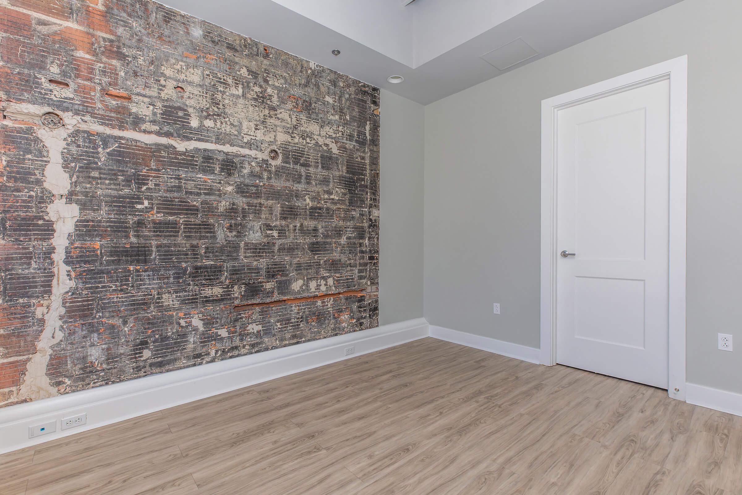 A minimalist room featuring a weathered brick accent wall and a door. The space has light-colored walls and a wooden floor, creating a modern and airy atmosphere. Natural light brightens the room, emphasizing the textural contrast between the rustic wall and the clean, contemporary design.