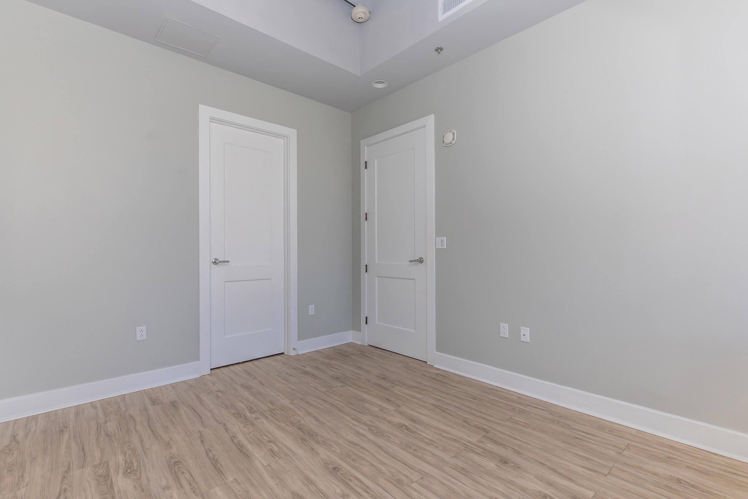 Empty room with light gray walls and wooden flooring. Two white doors are visible, one on the left and one on the right. Natural light comes from a window above, highlighting the spaciousness of the area. The ceiling is white and features a small vent.